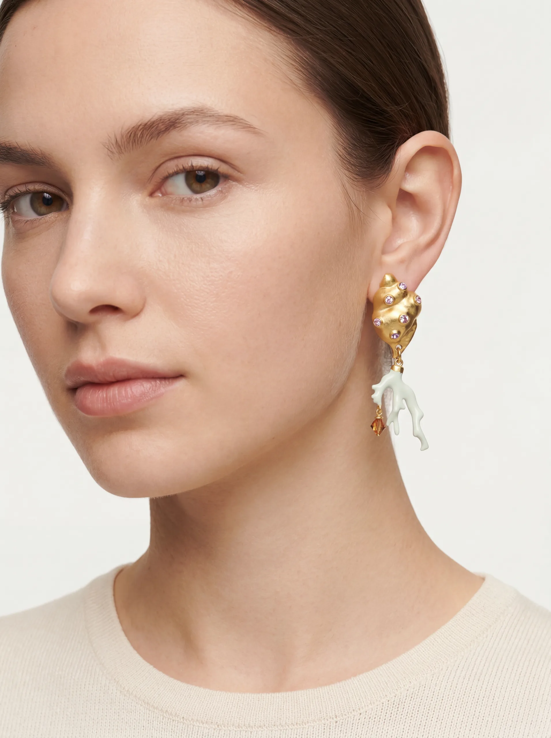 On-model close-up of a gold shell earring with pink crystals and a white coral drop. Oscar de la Renta AI Concept.