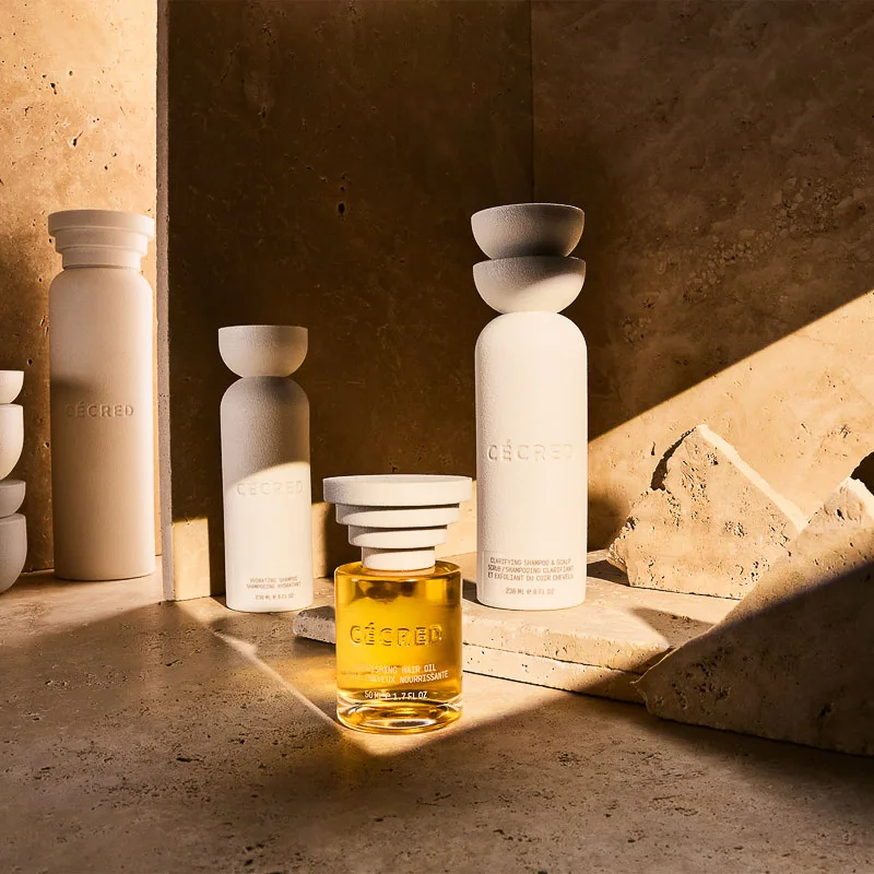 White hair care bottles and a golden hair oil on travertine stone in warm, angled sunlight, for Cécred.