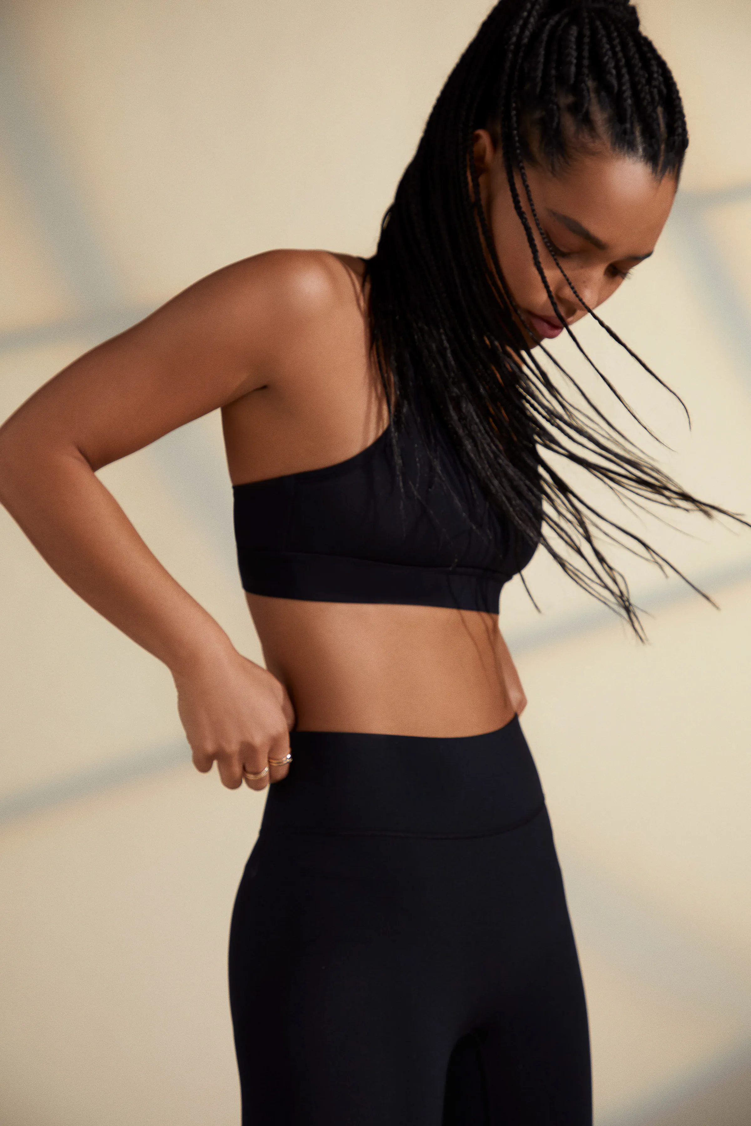 Model with long braids in a black sports bra and leggings against a warm beige wall with window shadows, for Bandier.