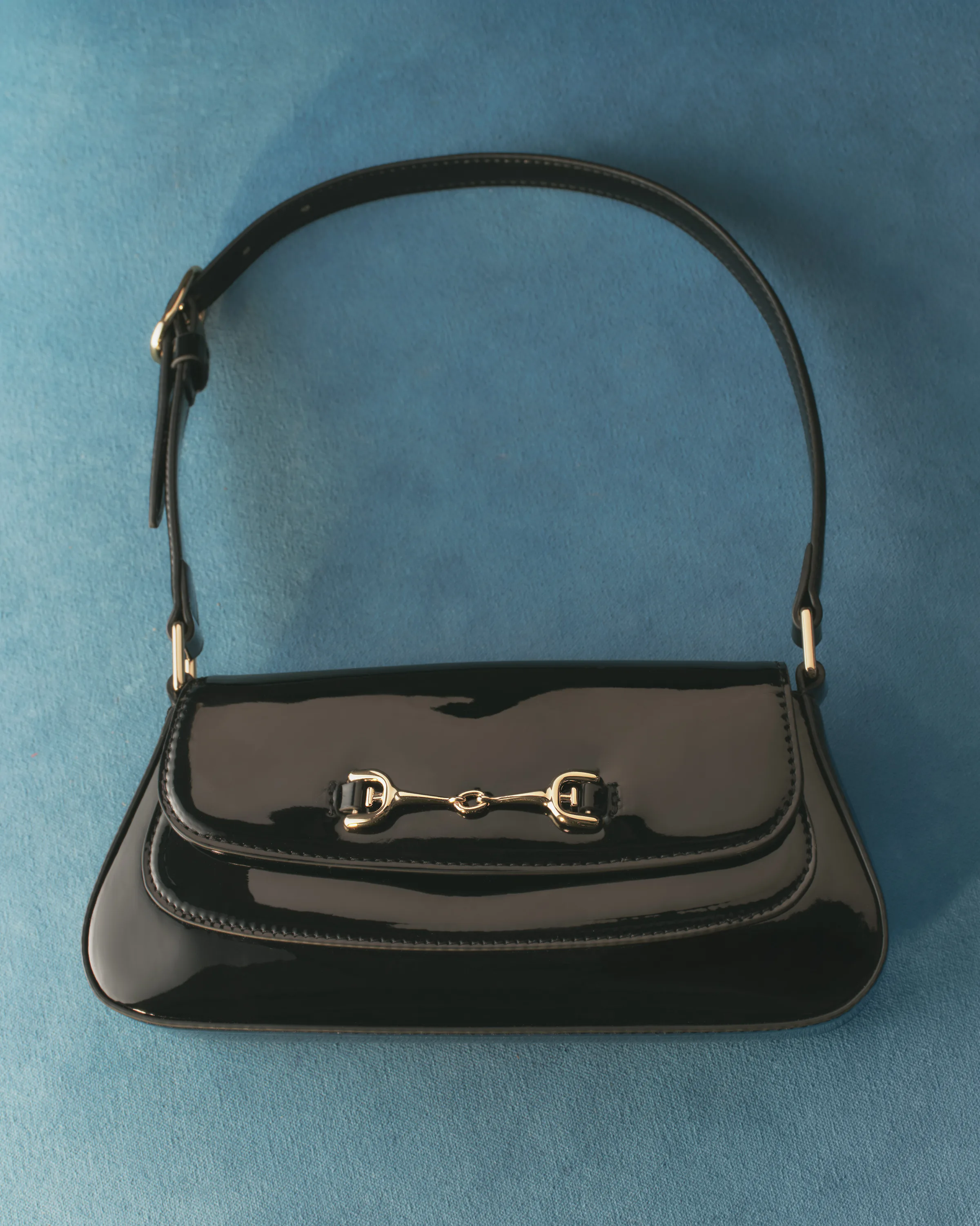 Black patent leather shoulder bag with gold horsebit hardware on a blue velvet backdrop, for Sam Edelman.