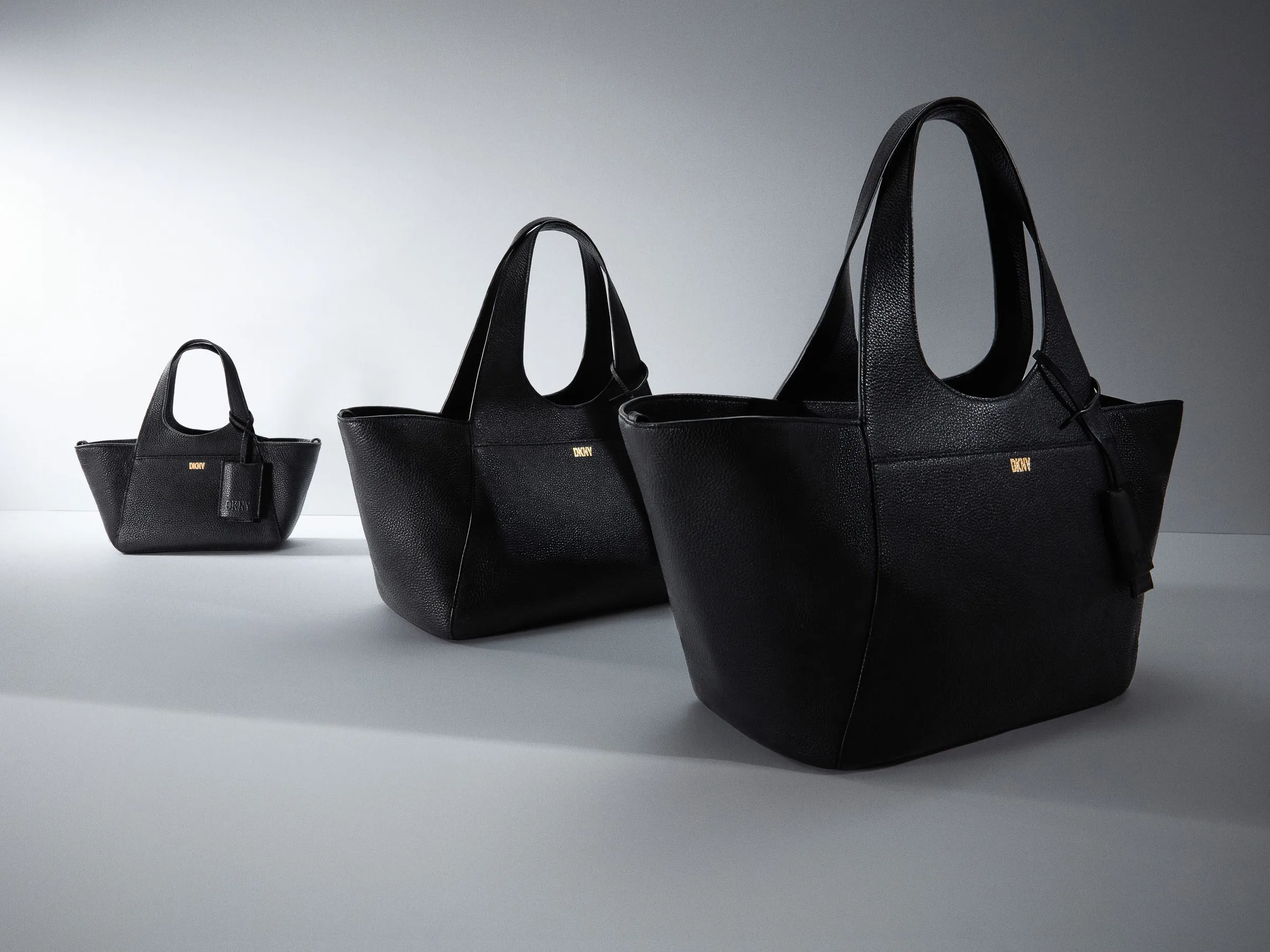 Three black pebbled leather tote bags with gold logo plaques, staggered small to large on a grey set, for DKNY.