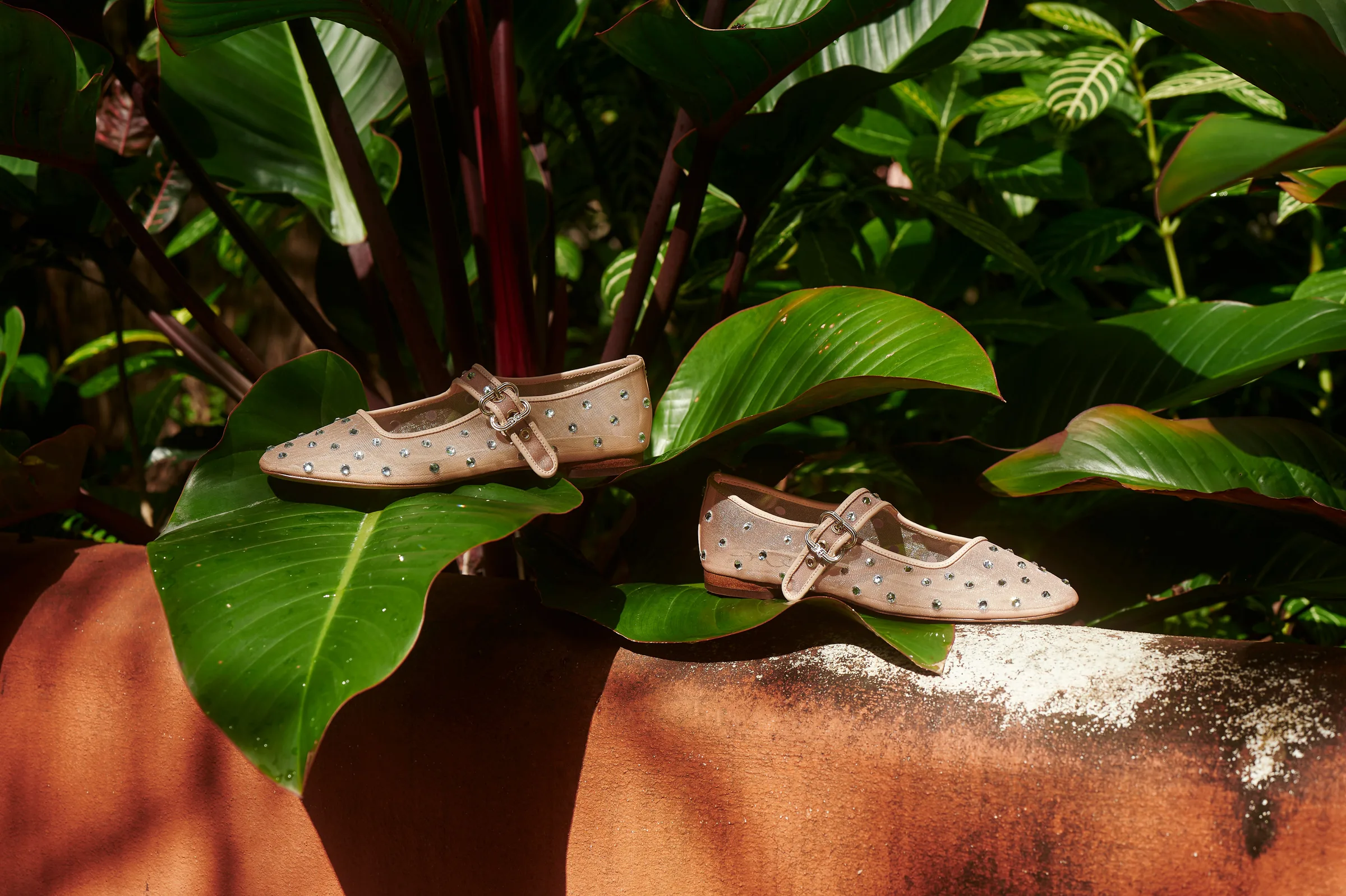Beige mesh Mary Jane flats with crystal studs resting on tropical leaves and a rust-red wall in sunlight, for Sam Edelman.