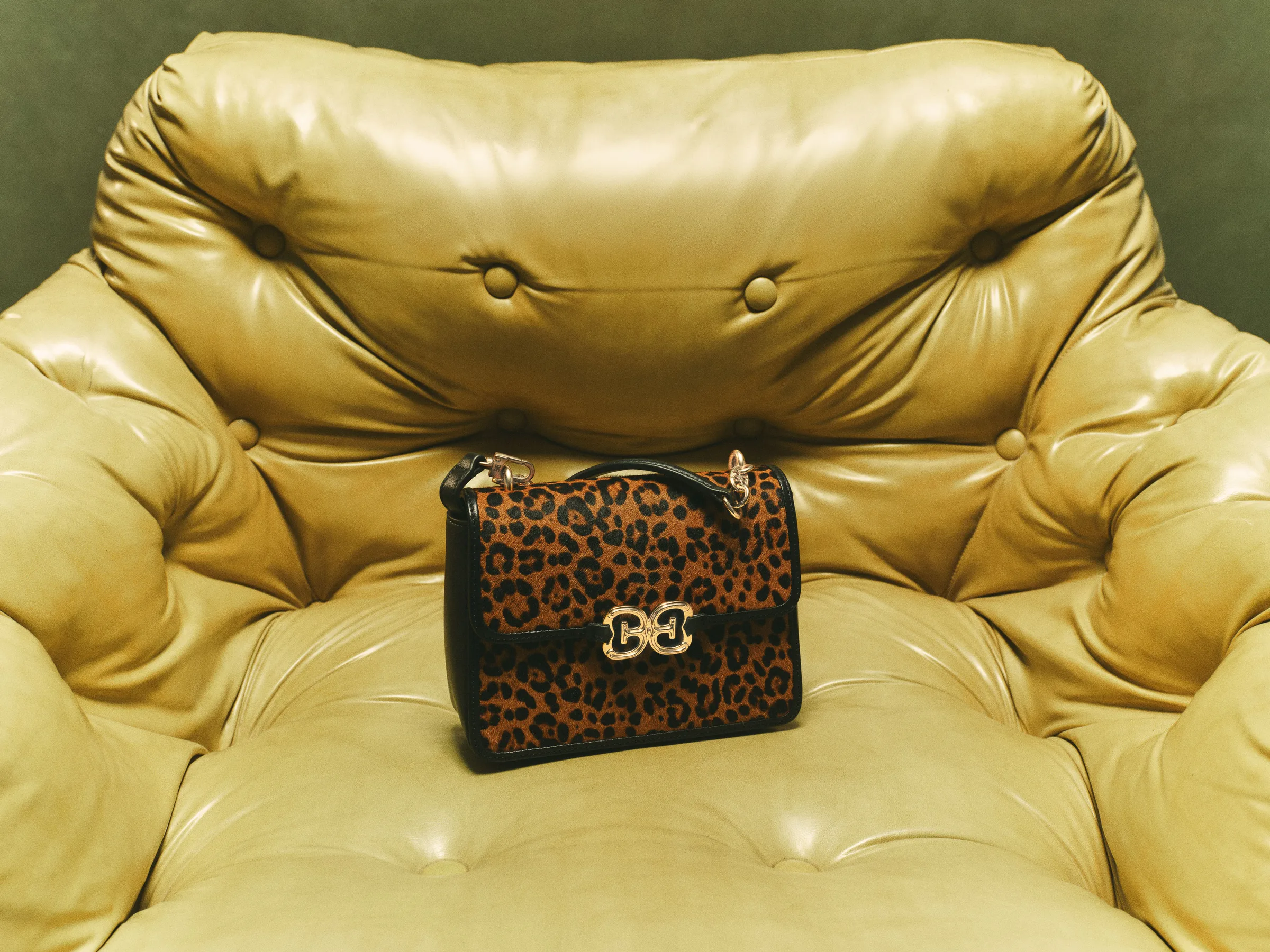 Leopard-print calf hair crossbody bag with gold double-E clasp on a tufted olive leather armchair, for Sam Edelman.