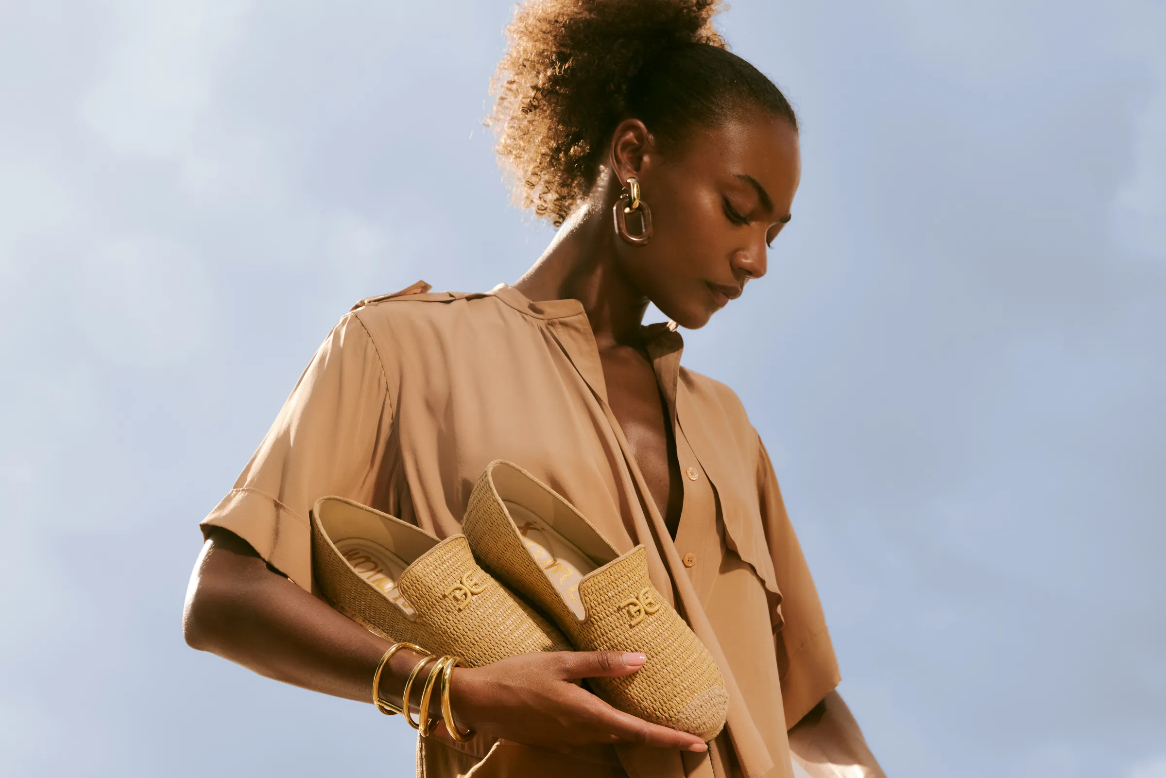 Model in a camel shirt dress holding woven raffia loafers with gold logo against a blue sky, for Sam Edelman.
