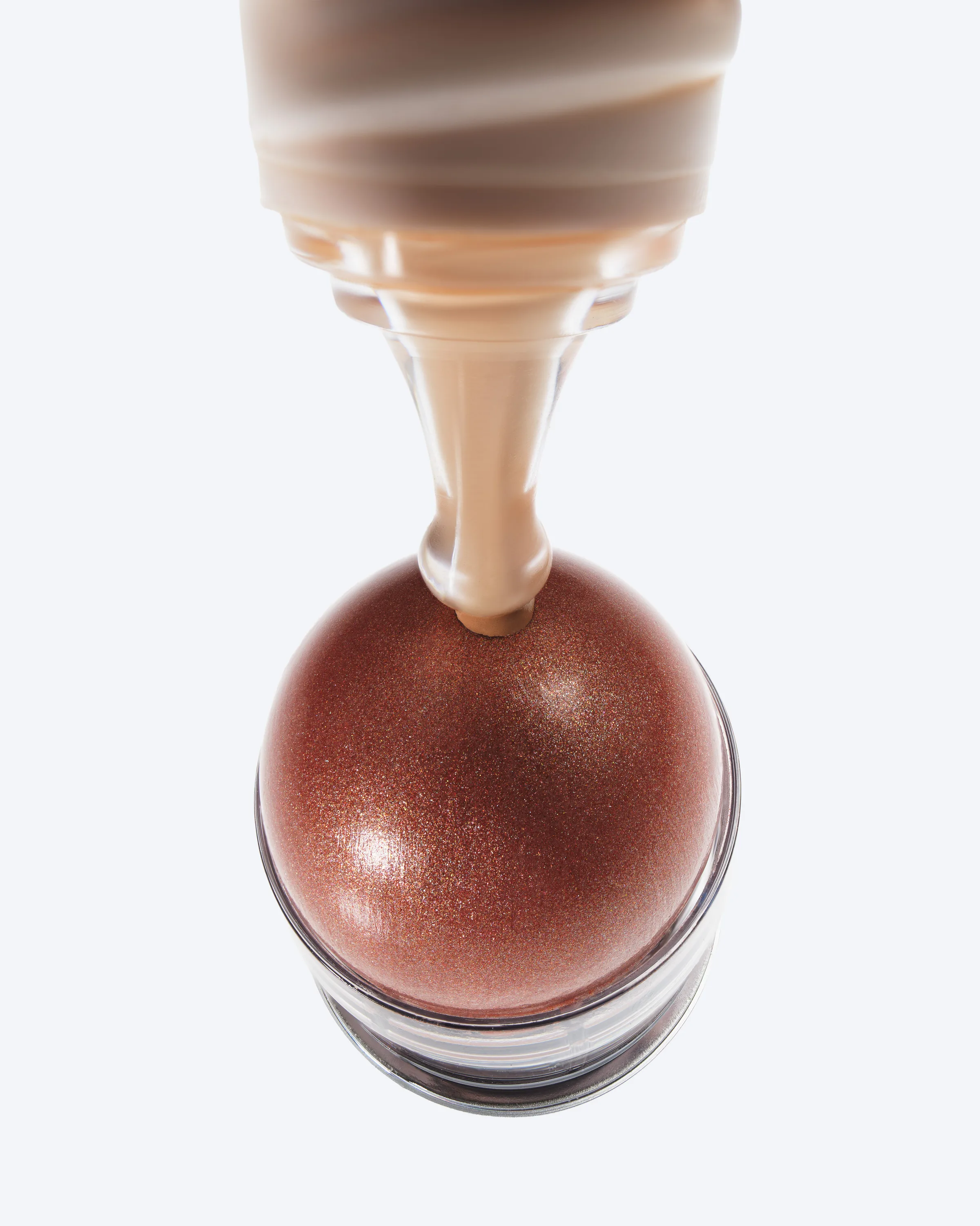 Peach skin tint pouring onto a shimmering bronze dewy balm stick against a pale grey background, for Kylie Cosmetics.