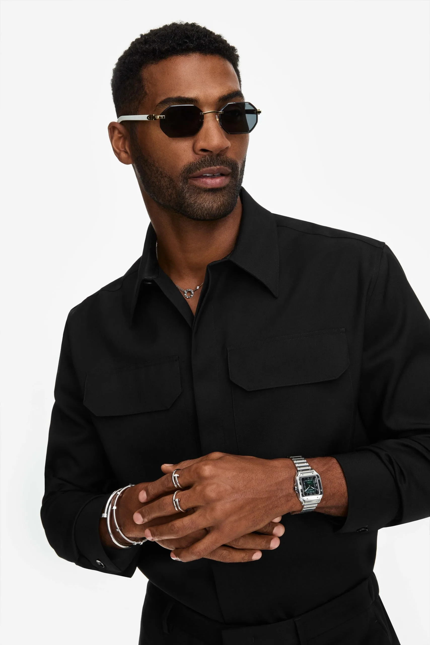 Man in black shirt and sunglasses wearing a steel watch, stacked rings and bracelets on white background, for Cartier.