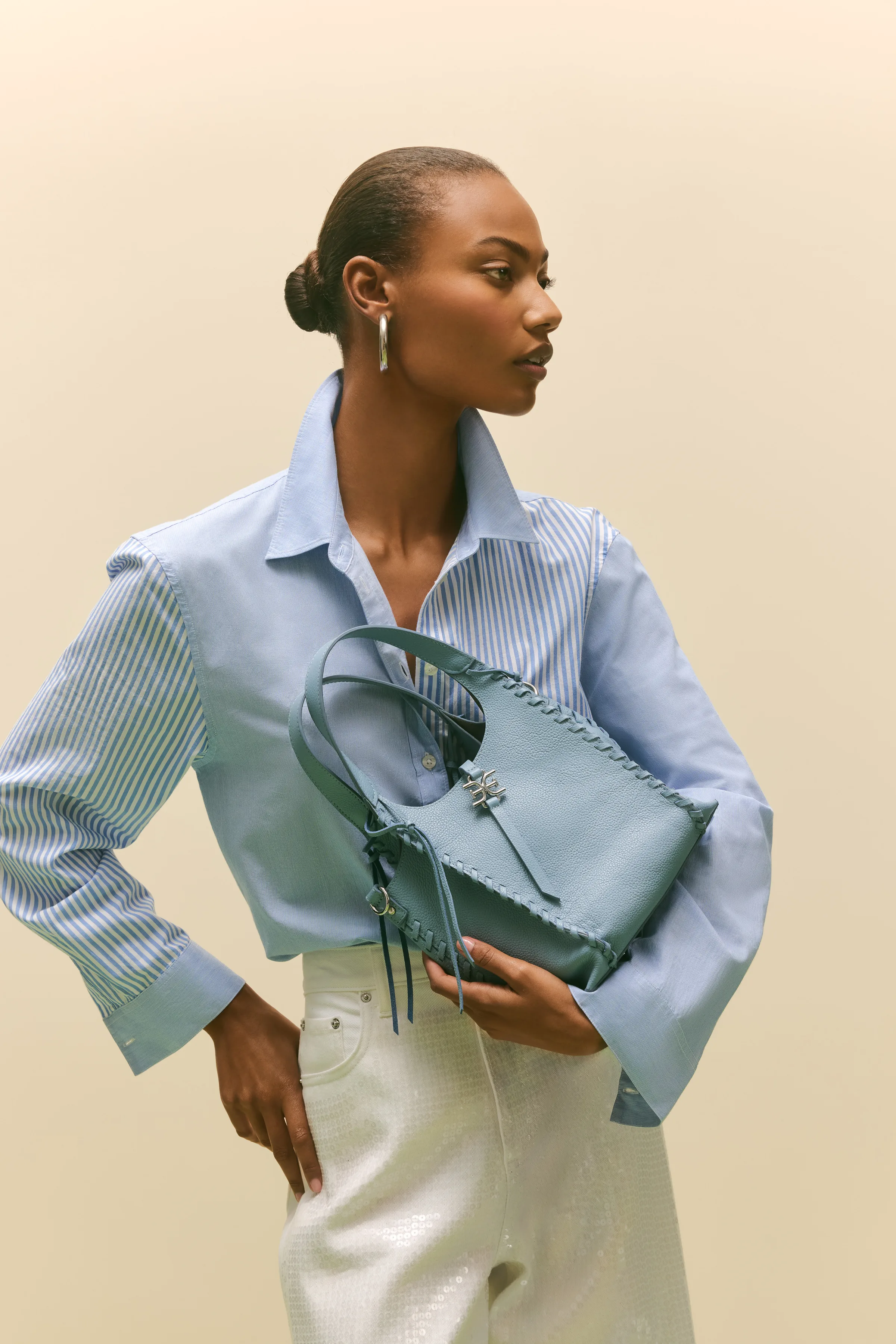 Model in a striped blue shirt and white sequin trousers holding a coastal blue whipstitched leather tote, for Sam Edelman.