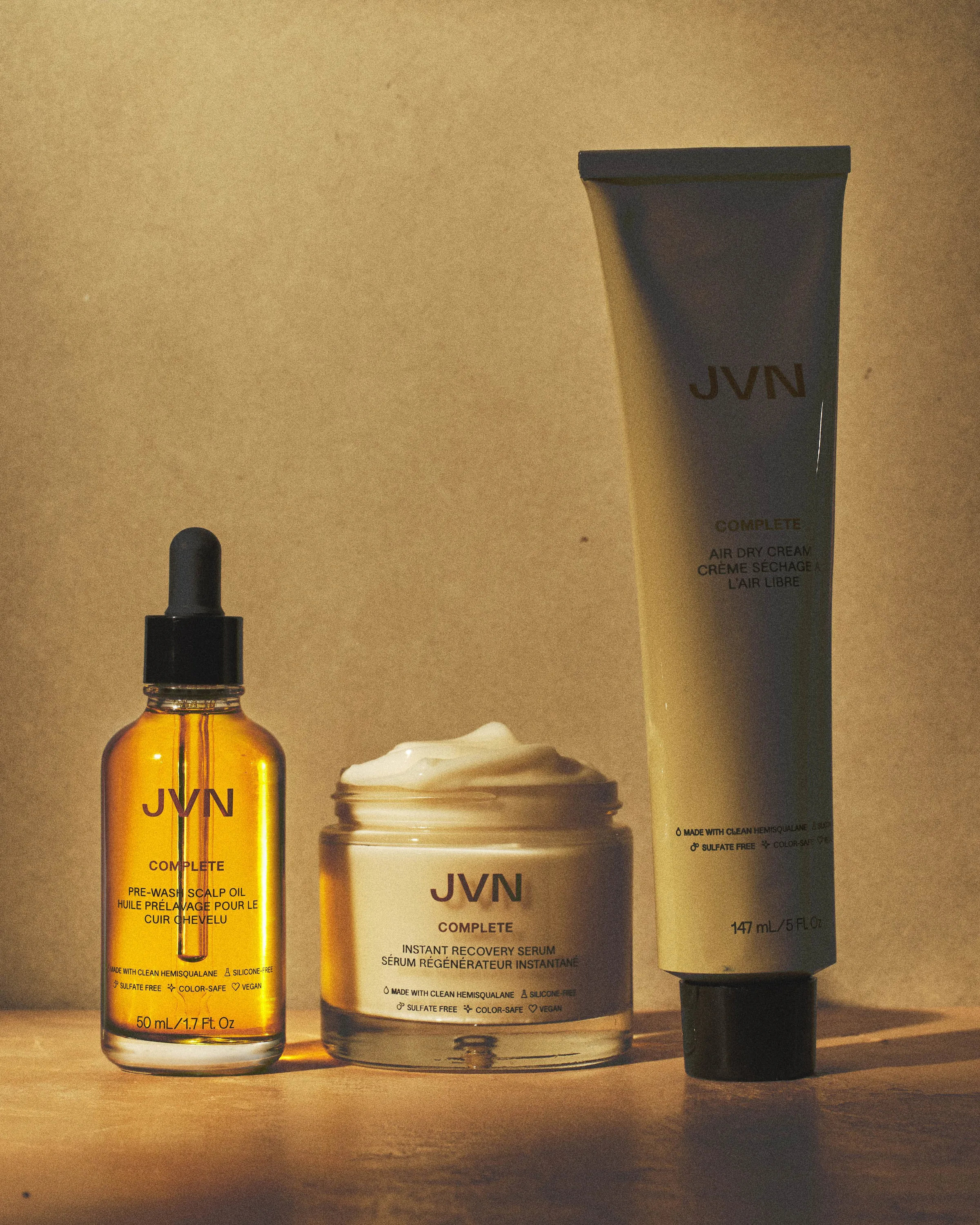 Hair care trio of scalp oil, open serum jar and air dry cream tube in warm side light on a beige backdrop, for JVN.