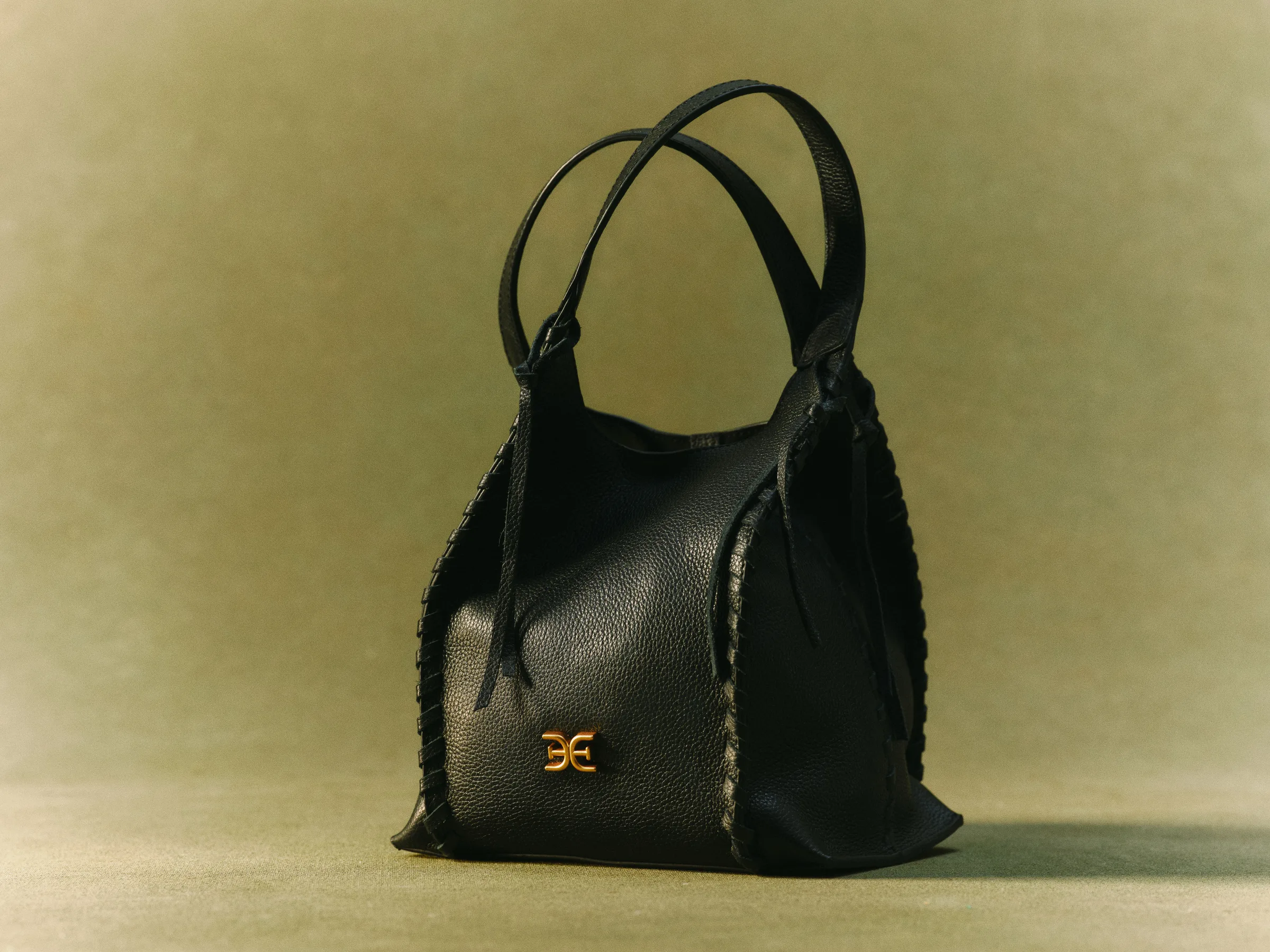 Black pebbled leather tote with whipstitched sides and gold logo hardware on an olive green backdrop, for Sam Edelman.