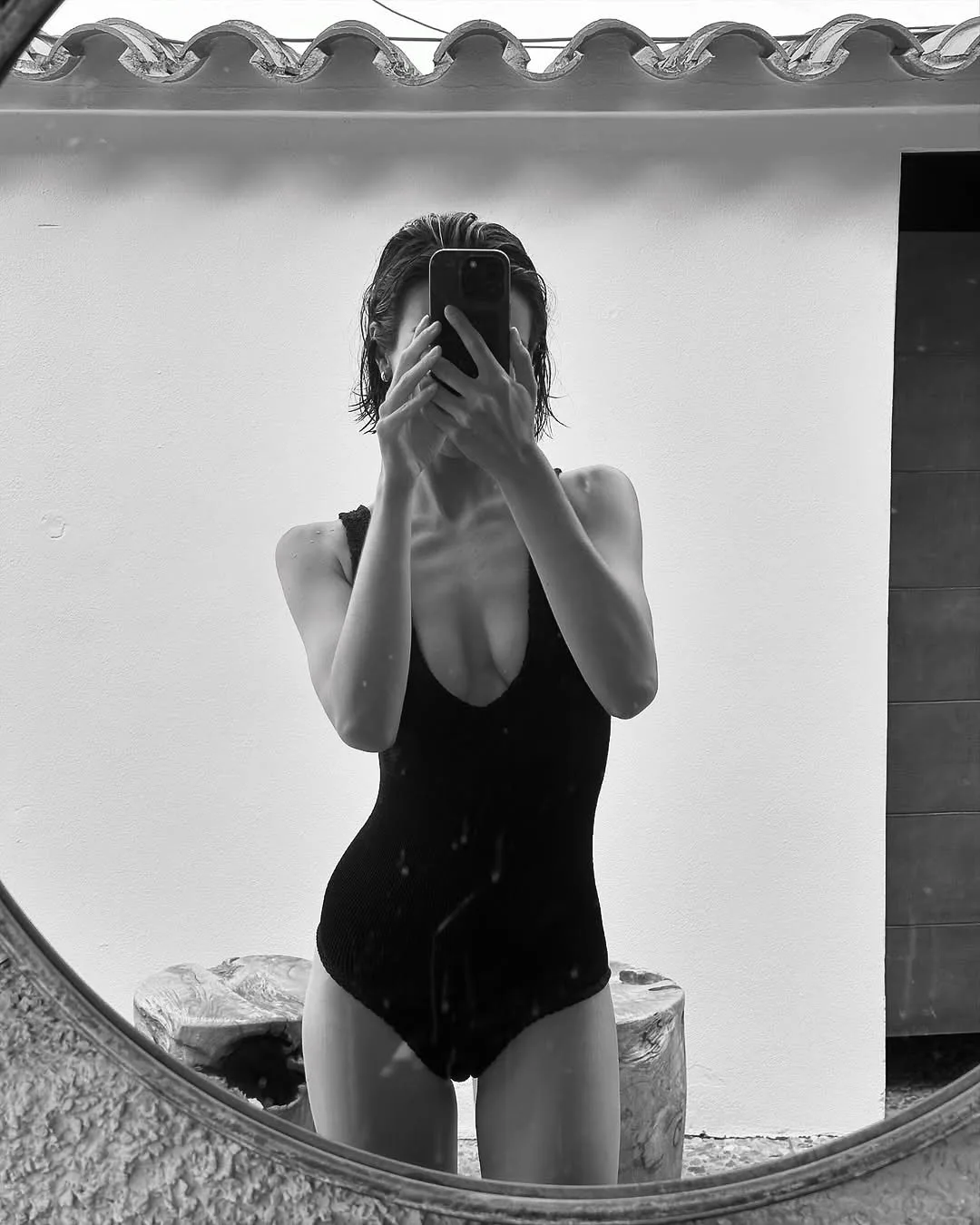 Black-and-white mirror selfie of a woman with wet hair in a black ribbed one-piece swimsuit, phone covering her face.