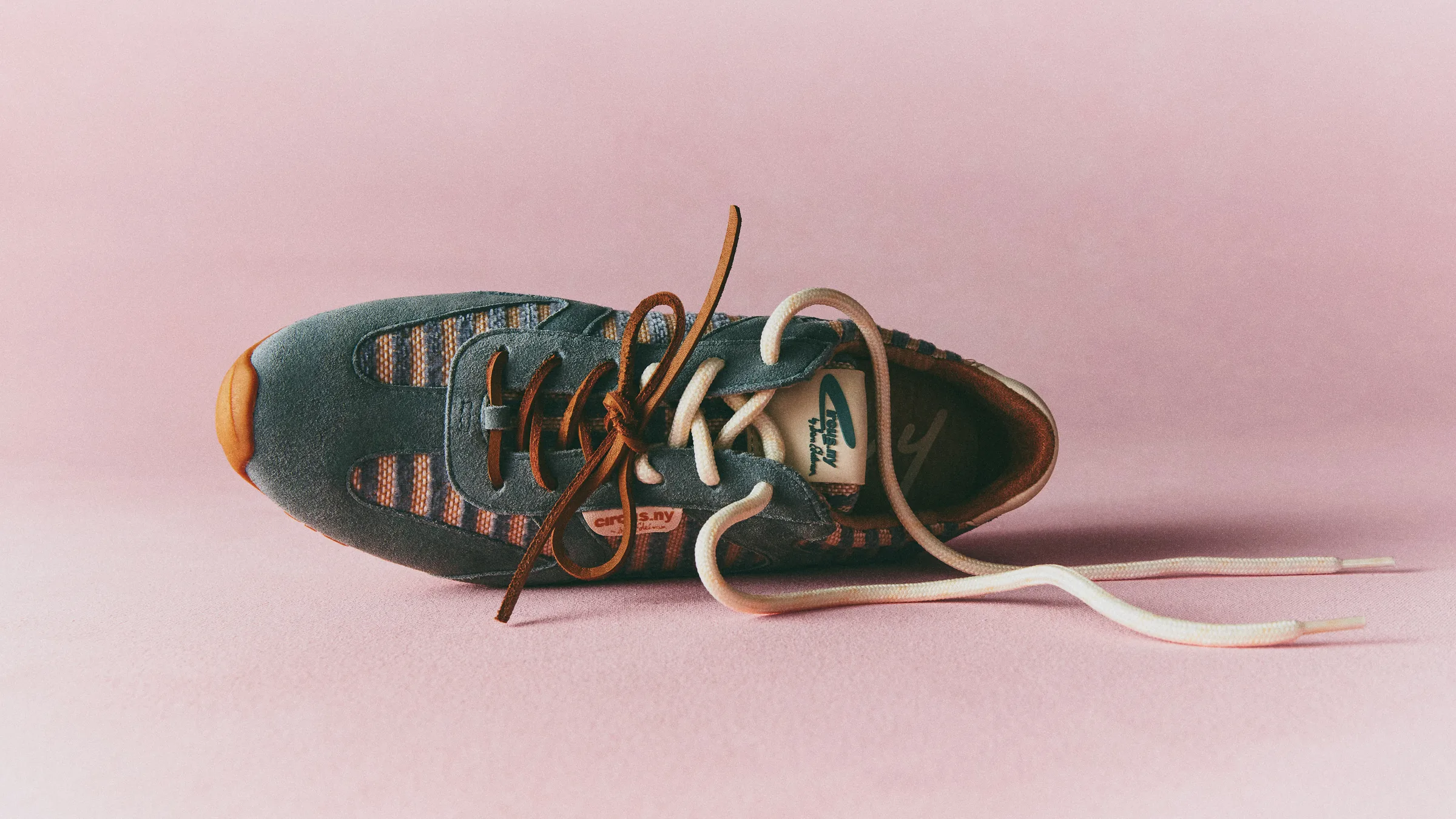 Blue suede Nash sneaker with striped panels and tan and cream laces on a pink backdrop, for Circus NY.