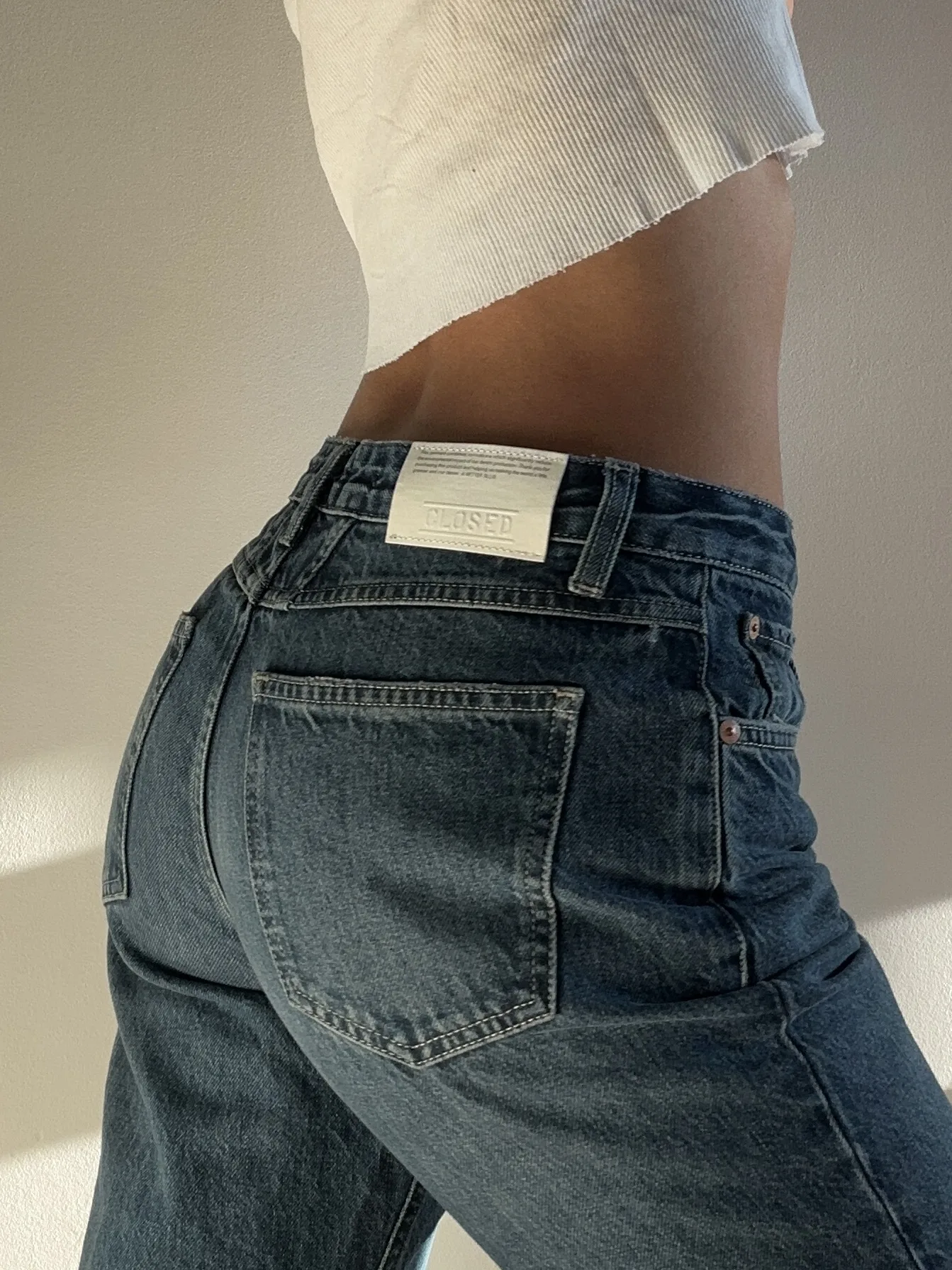 Back view of a model in dark wash straight jeans with a leather patch and white cropped knit top, in soft sunlight, for Closed.