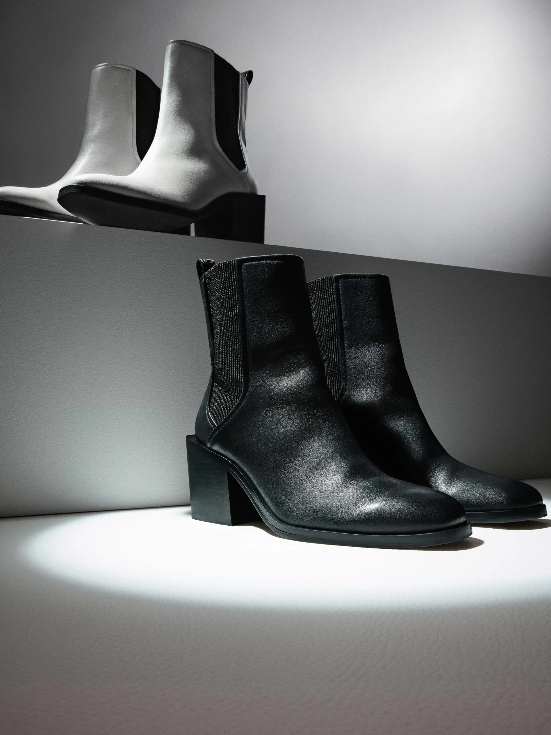Black leather block-heel Chelsea boots under a spotlight, white pair on a step behind, for DKNY.