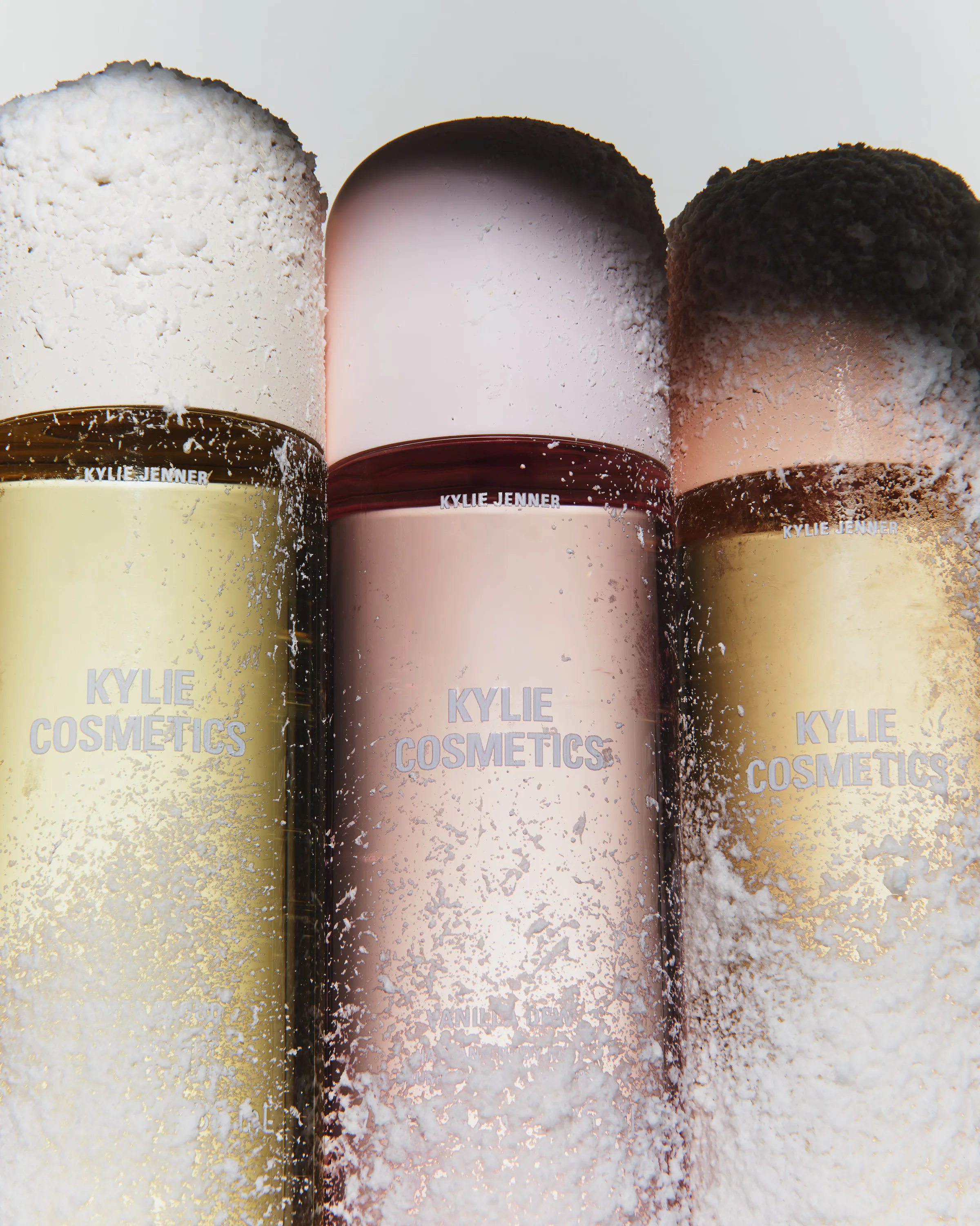 Three gold and pink body mist bottles dusted with artificial snow in a close-up still life, for Kylie Cosmetics.