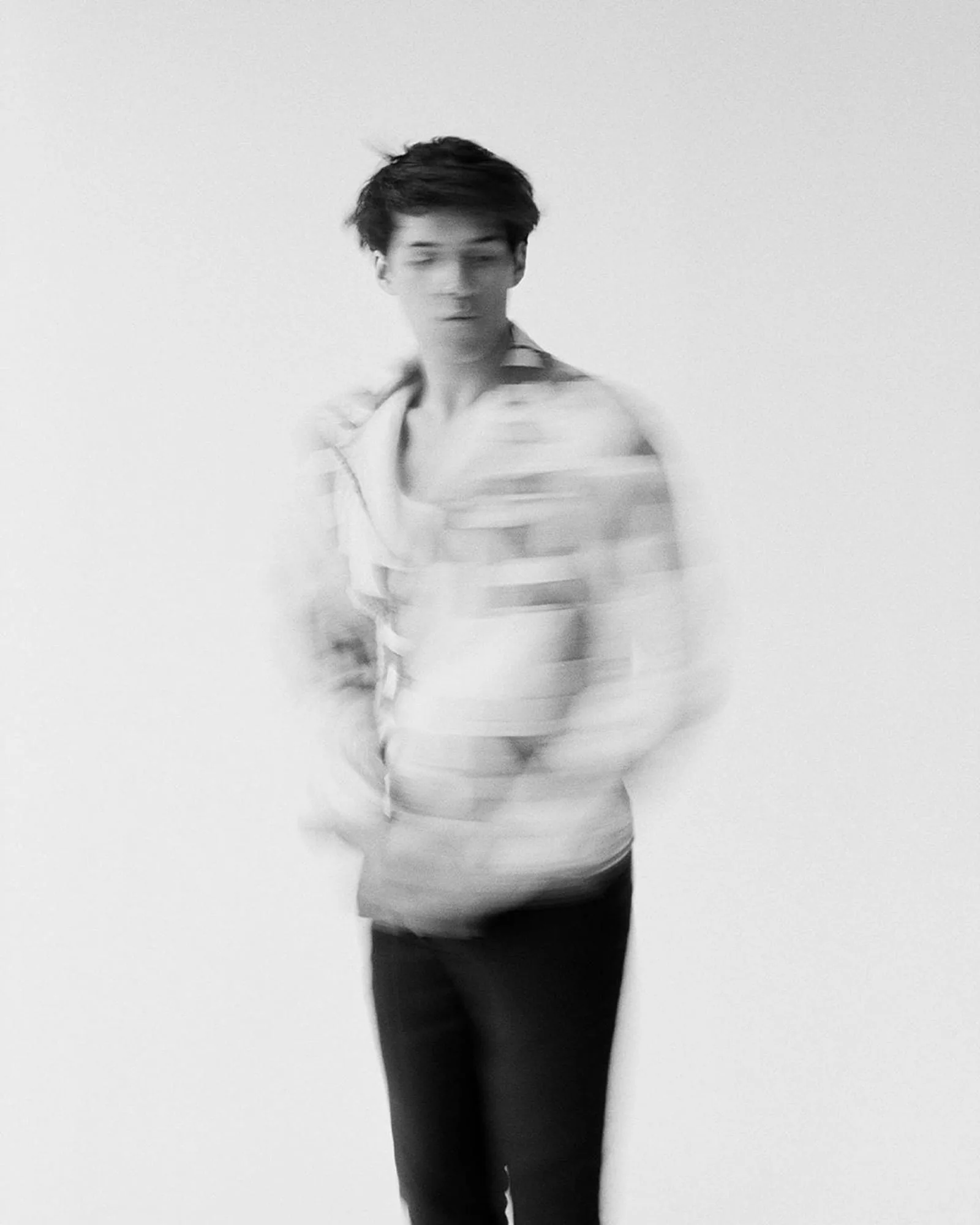 Black and white motion-blurred portrait of a dark-haired man in a striped shirt against a white wall, for The Last Magazine.
