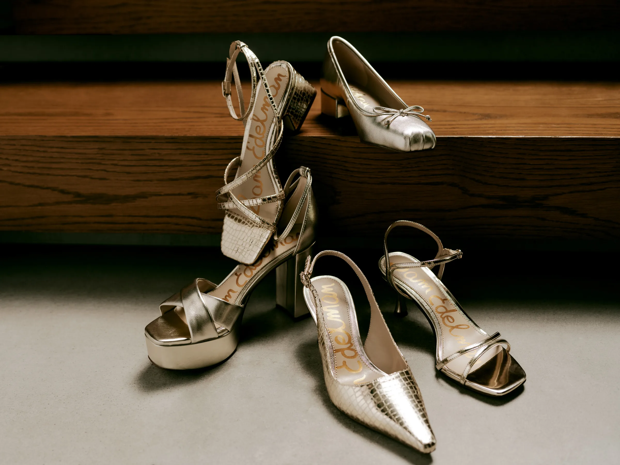 Five metallic gold and silver heels, sandals and flats arranged on wooden steps and a grey floor, for Sam Edelman.