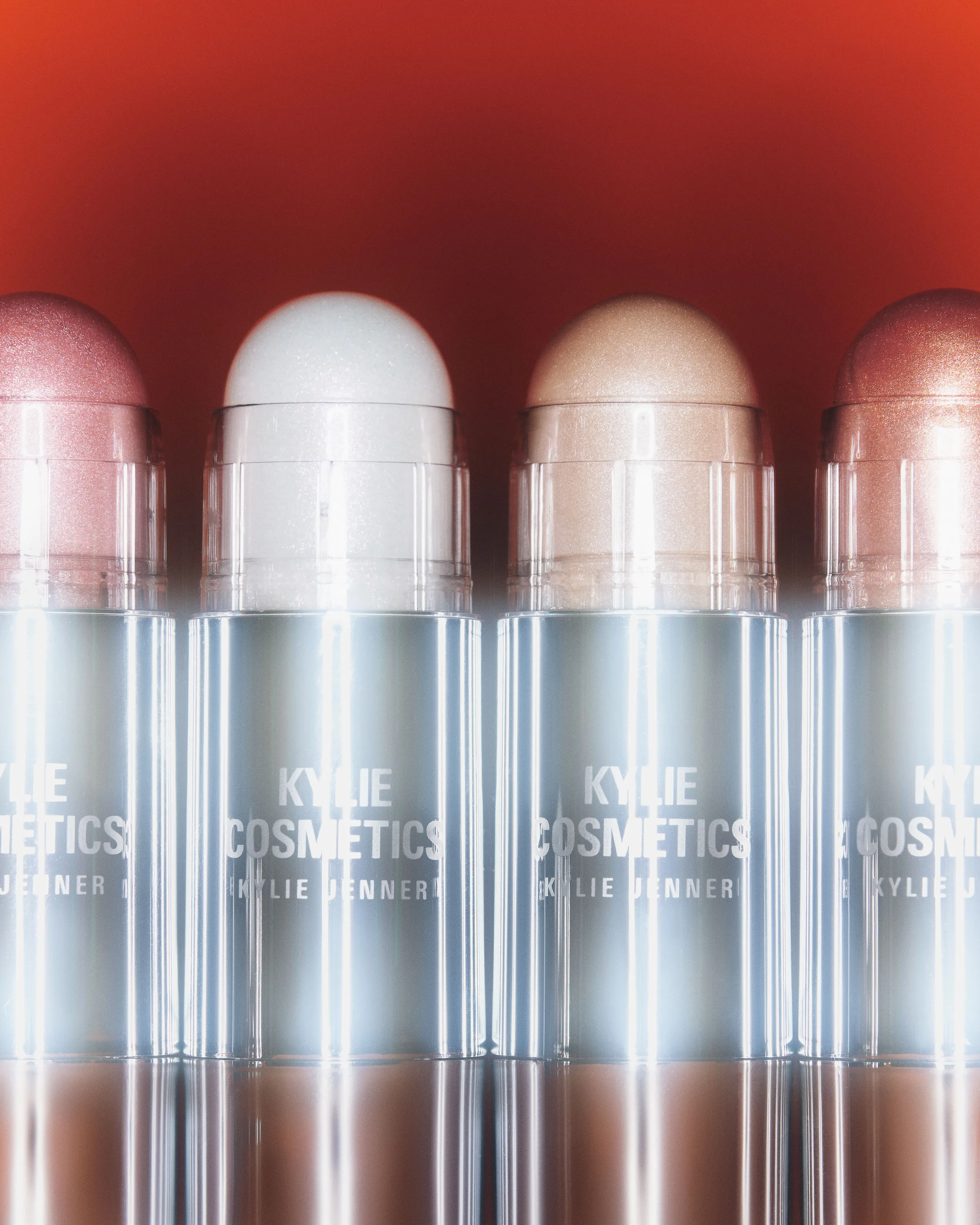 Row of shimmering dewy balm sticks in pink, white, champagne and rose shades against an orange backdrop, for Kylie Cosmetics.