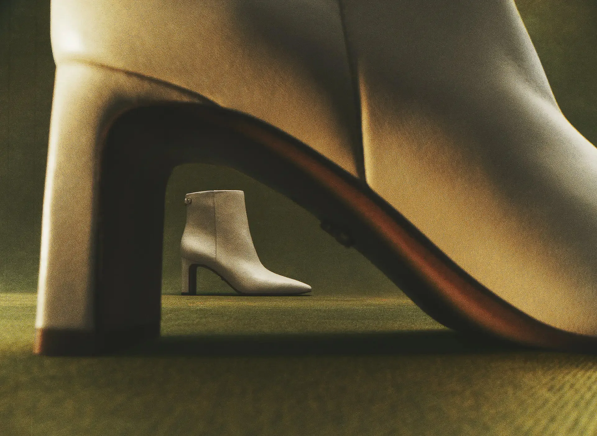 Small cream ankle boot framed beneath the arch of a giant cream heeled boot on green carpet, for Sam Edelman.