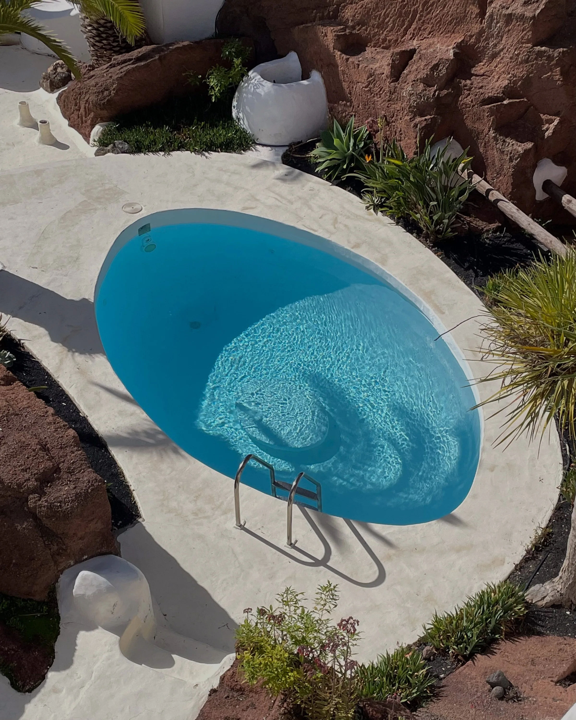 Overhead view of an oval blue pool with a steel ladder, white stone deck, red volcanic rock and desert plants.