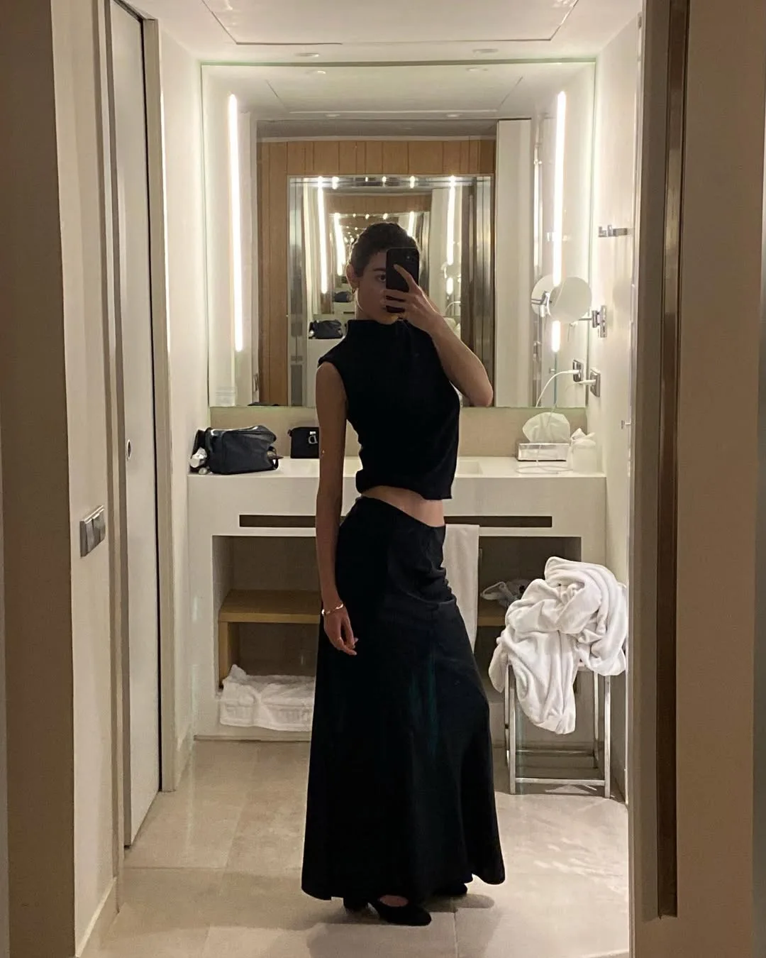 Woman takes a mirror selfie in a hotel bathroom wearing a black sleeveless top and long black maxi skirt.
