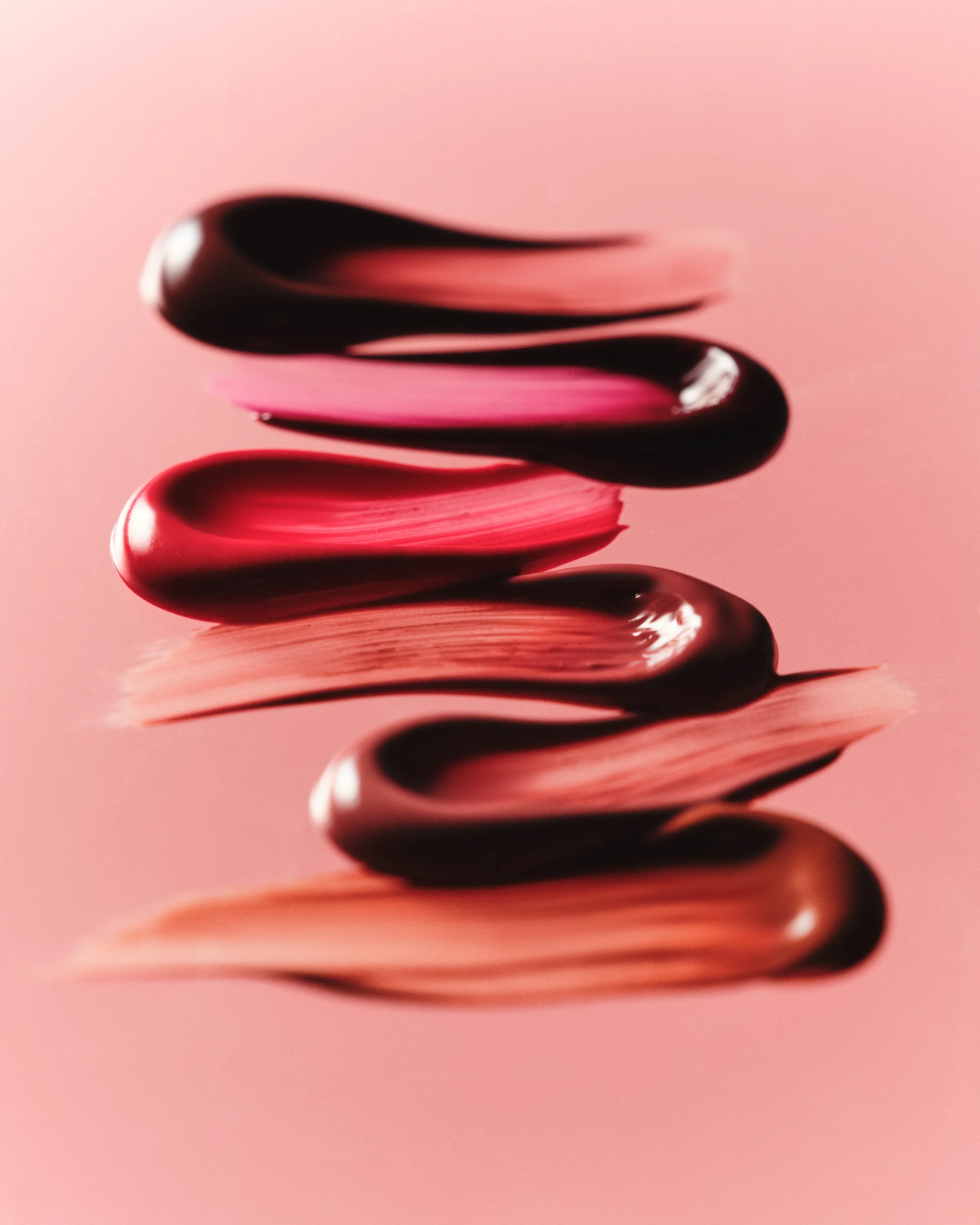 Stacked glossy lip product swatches in berry, red and nude shades smeared across a pink background, for Kylie Cosmetics.