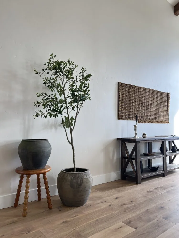 Potted olive tree beside a bobbin stool and dark wood console under a woven wall hanging on oak floors, for Jennifer Hagler.