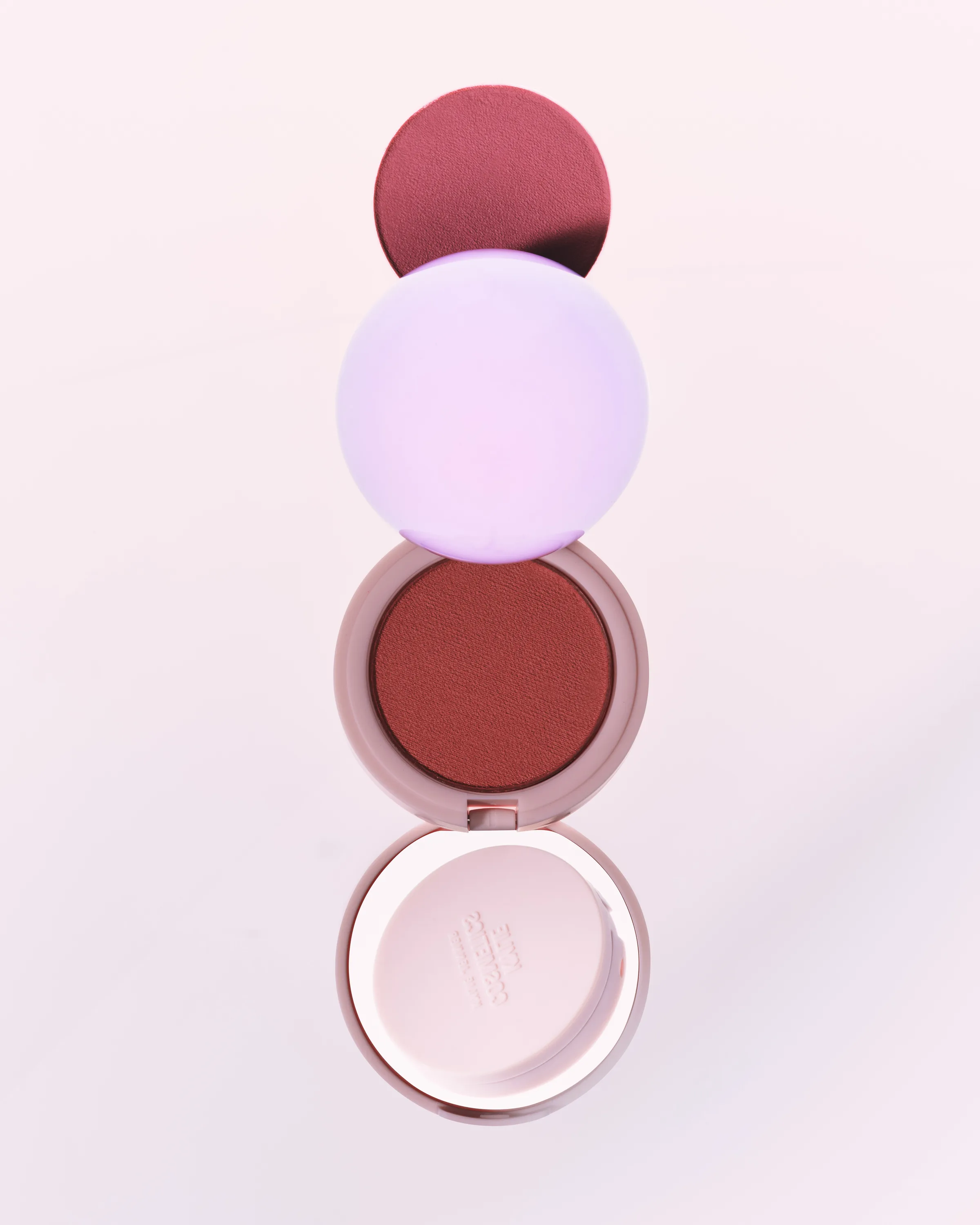 Open blush compact with berry-red powder, sponge and lilac sphere stacked vertically on a pale pink background, for Kylie Cosmetics.