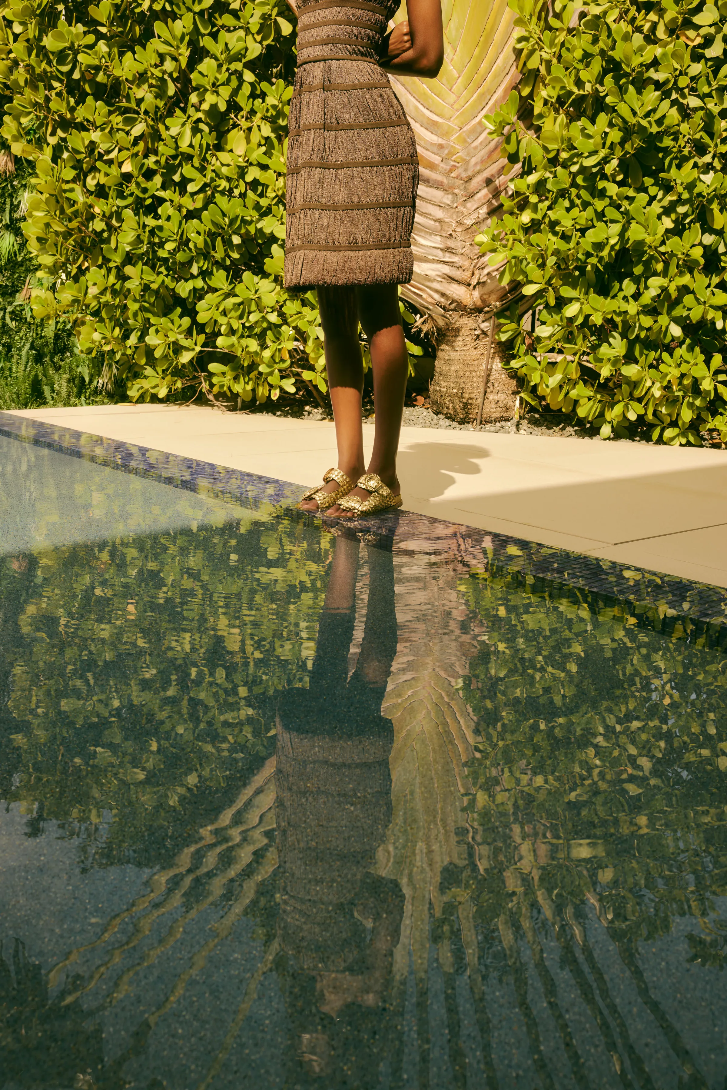 Model in a fringed brown dress and gold woven sandals stands at a pool edge, reflected in the water, for Sam Edelman.