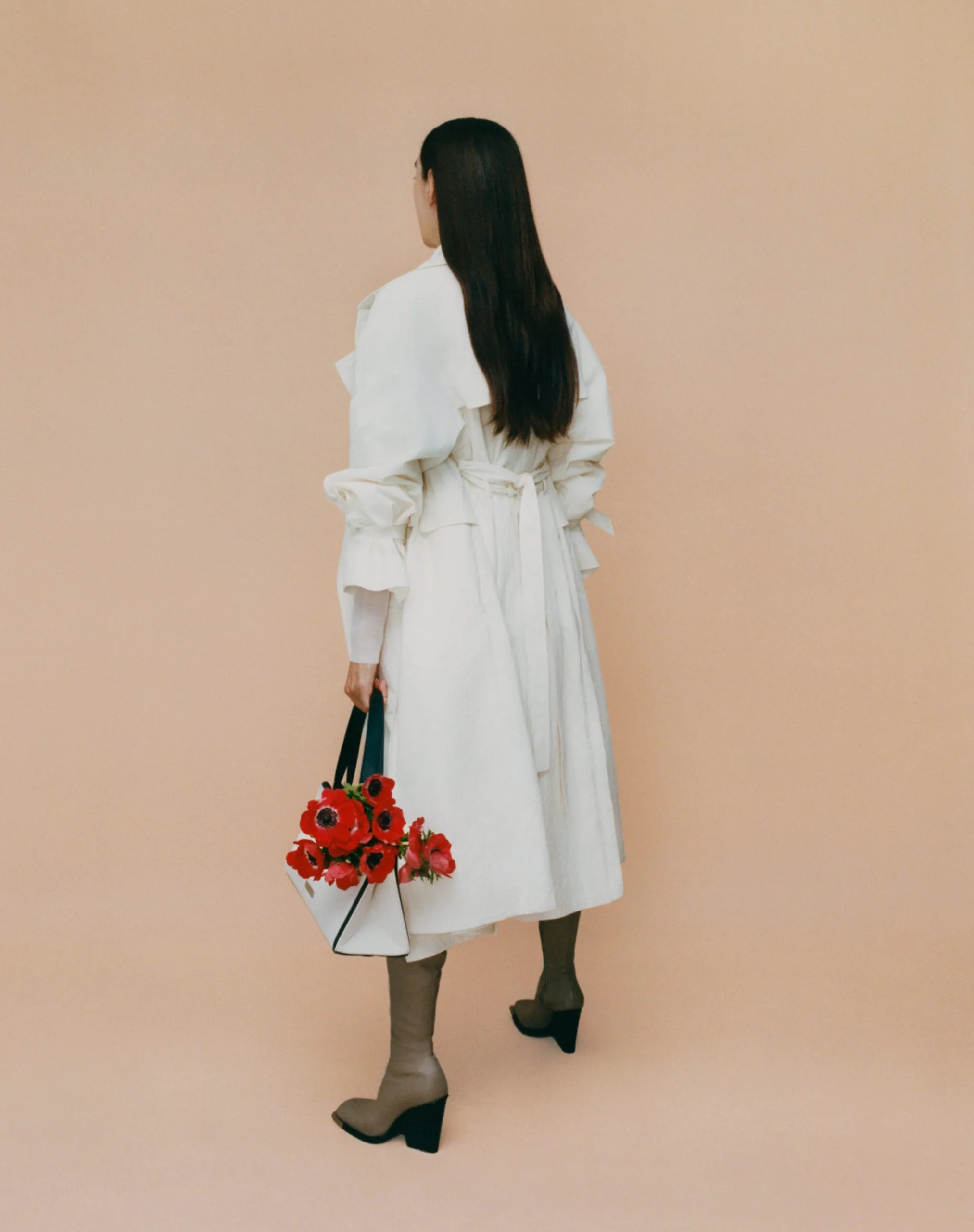 Model seen from behind in a cream ruffled trench coat and taupe boots, holding a tote of red anemones, for Interview Magazine.
