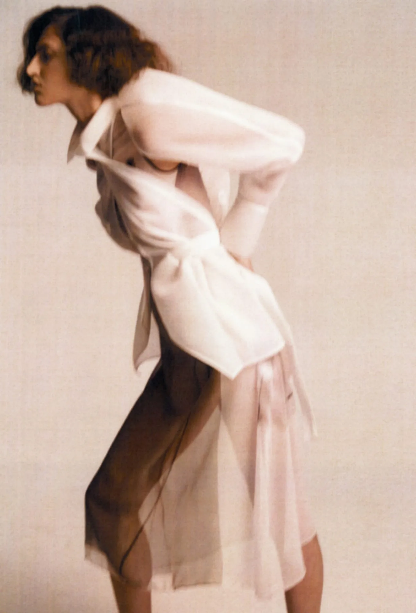 Model leaning forward in a sheer white organza trench dress against a warm beige backdrop, shot by Amber Witcomb.