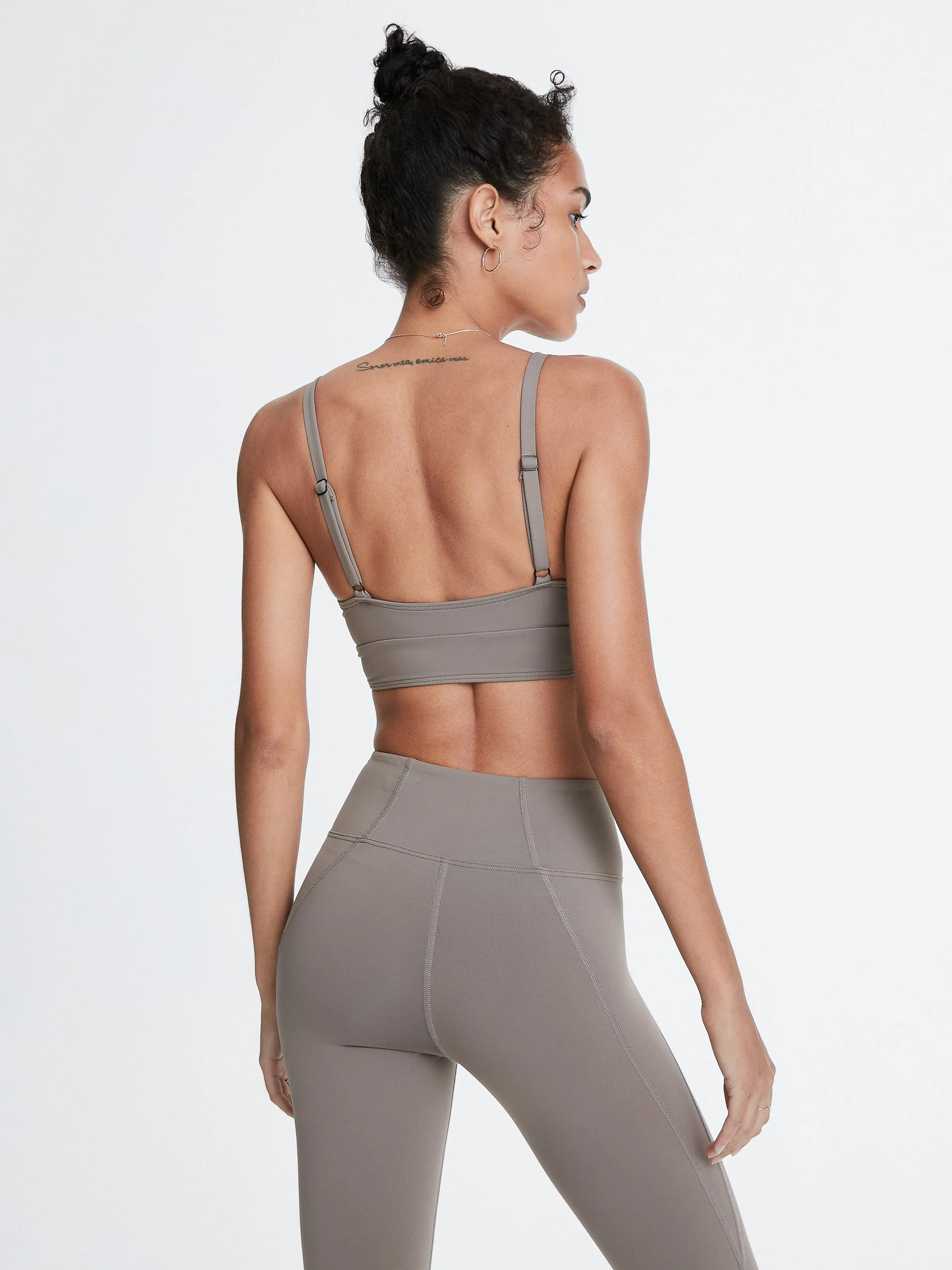 Model from behind in a taupe strappy sports bra and matching leggings on a white background, for Bandier.