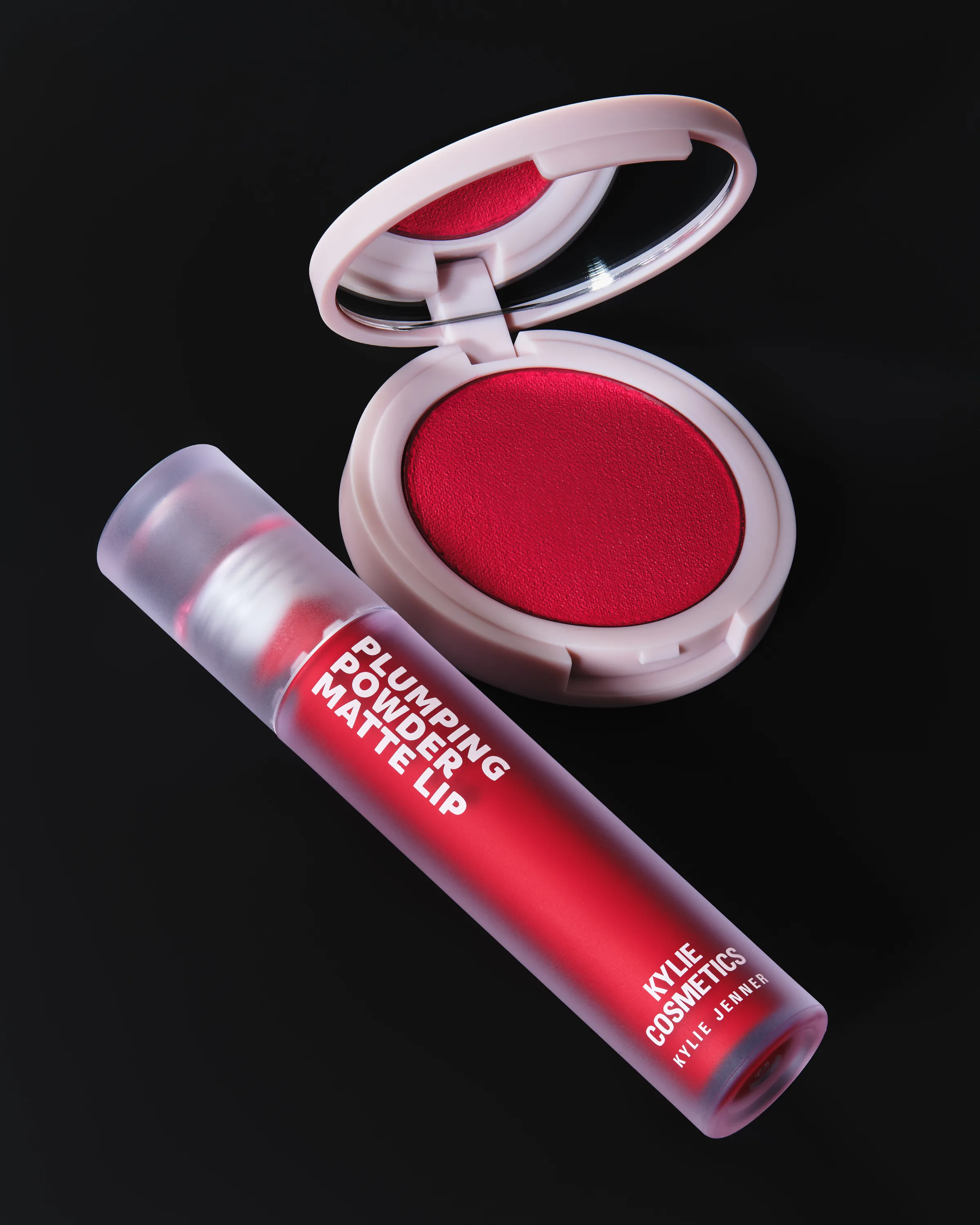 Still life of a red plumping powder matte lip tube and open blush compact with mirror on black, for Kylie Cosmetics.