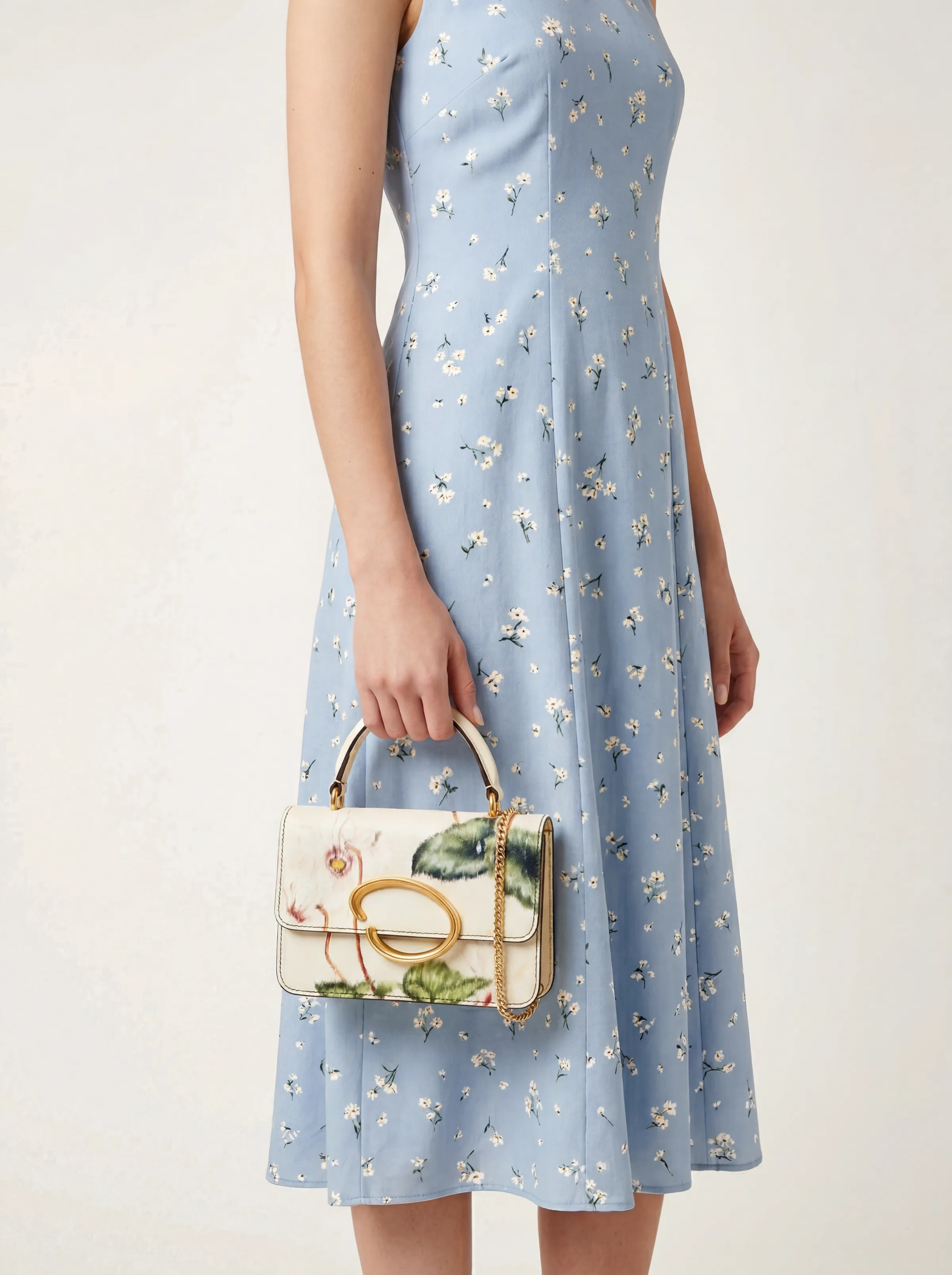 On-model shot of a cream floral top-handle bag with gold O clasp, held against a blue daisy-print dress. Oscar de la Renta AI Concept.
