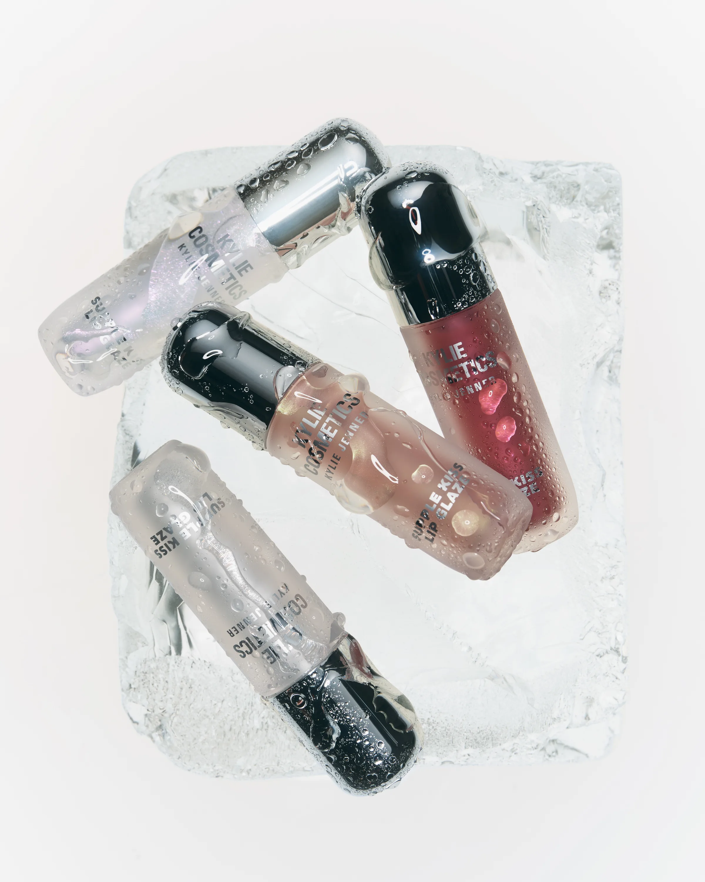 Four lip glaze tubes in clear, pink and berry shades with water droplets resting on a block of ice, for Kylie Cosmetics.