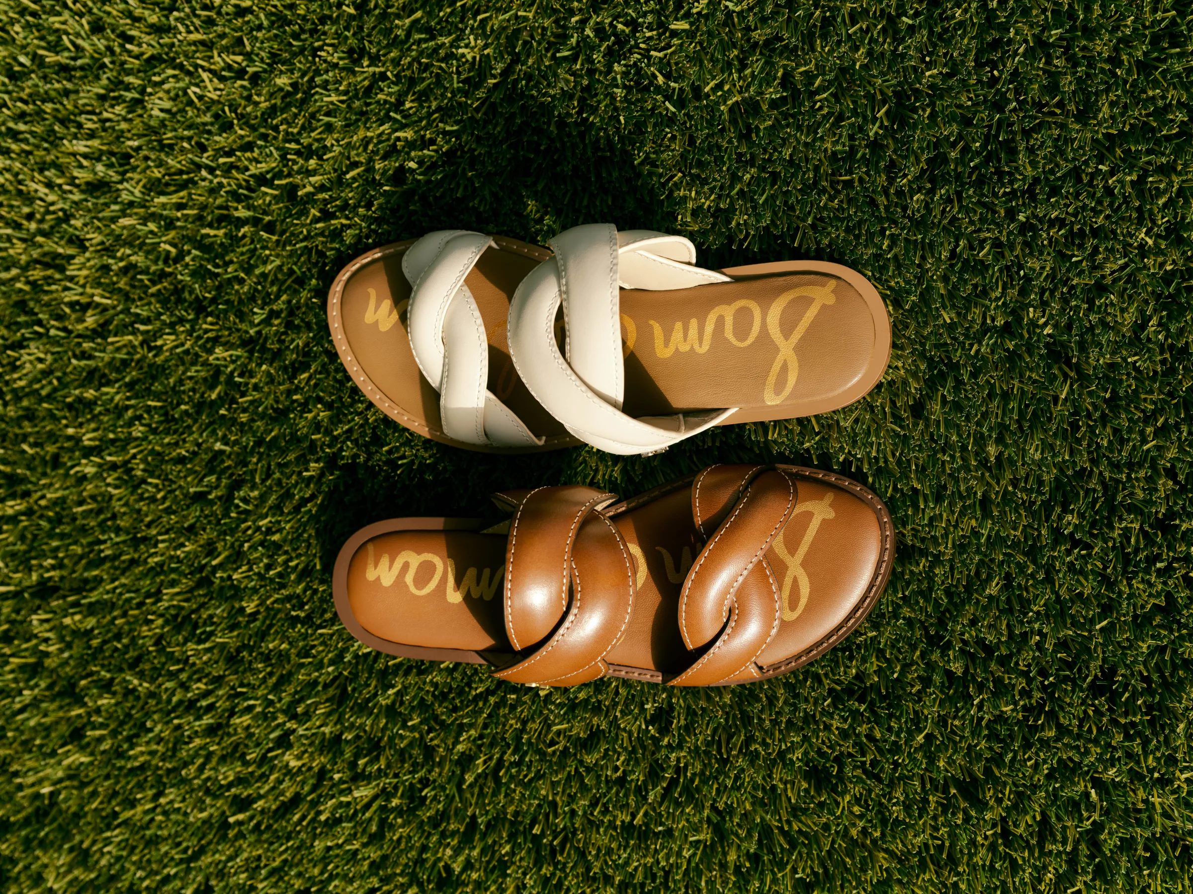 Cream and cognac leather slide sandals with twisted straps laid on green grass in sunlight, for Sam Edelman.