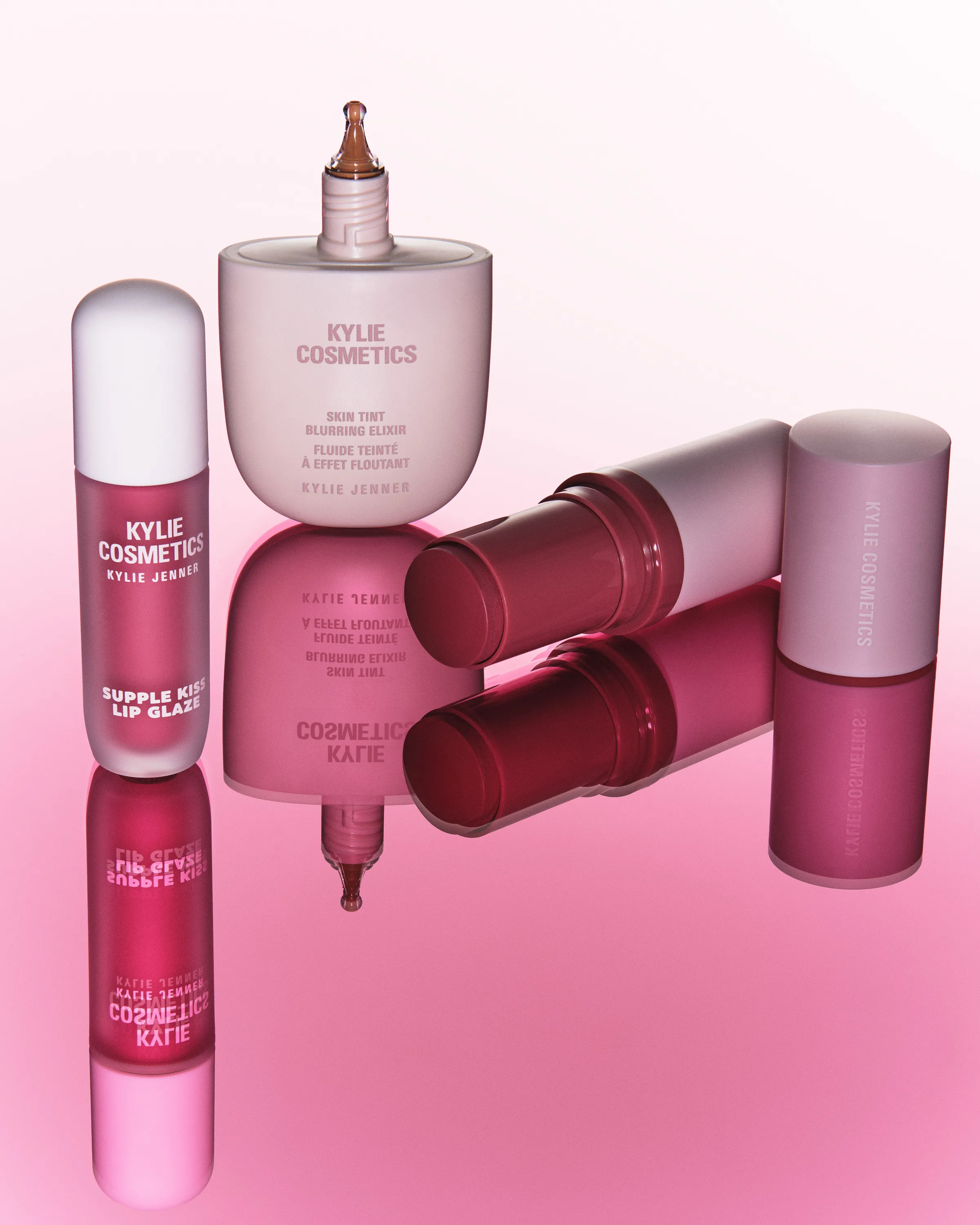 Lip glaze, skin tint, blush stick and lip oil reflected on a glossy pink surface, for Kylie Cosmetics.