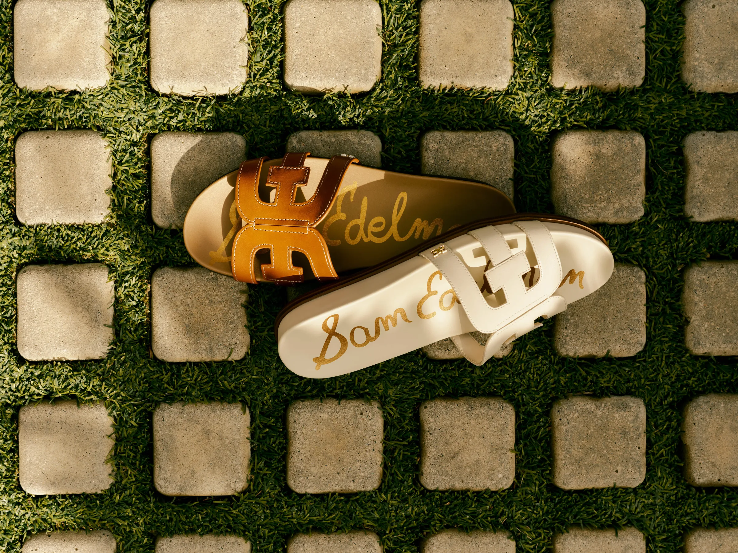 Tan and cream leather slide sandals with logo straps on grass between stone pavers, for Sam Edelman.