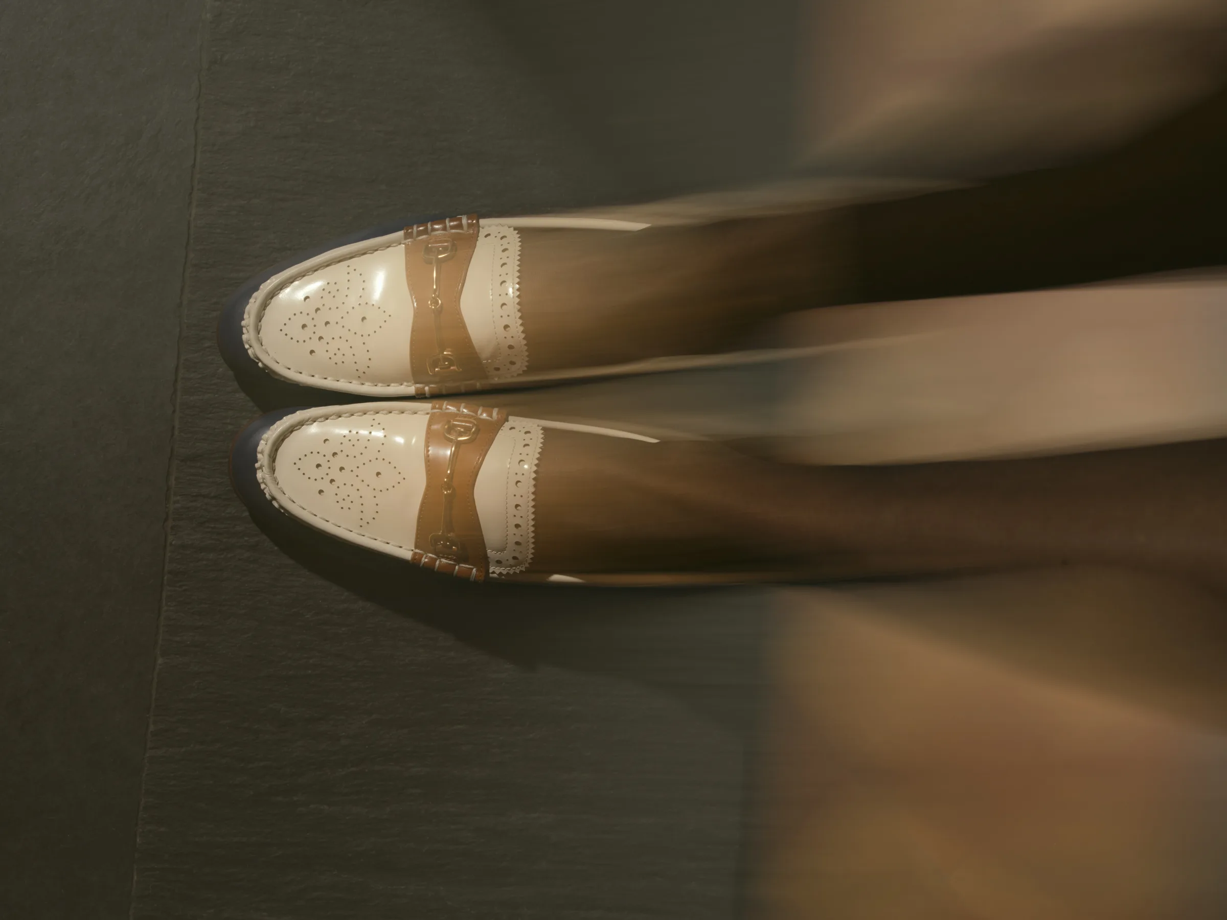 Cream and tan patent brogue loafers with horsebit detail worn on grey carpet, legs blurred in motion, for Sam Edelman.