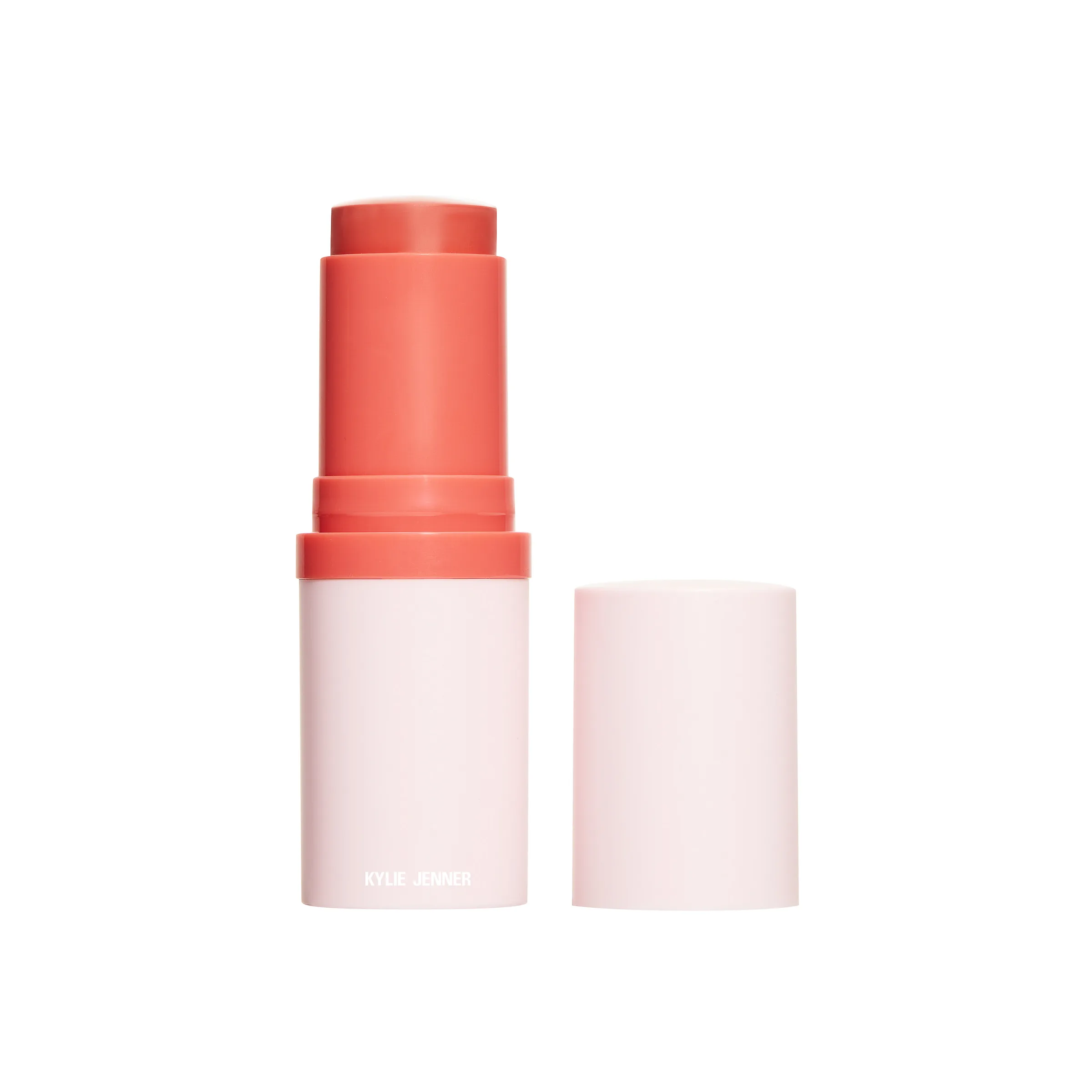 Coral cream blush stick uncapped in a pale pink tube beside its lid on white, for Kylie Cosmetics.