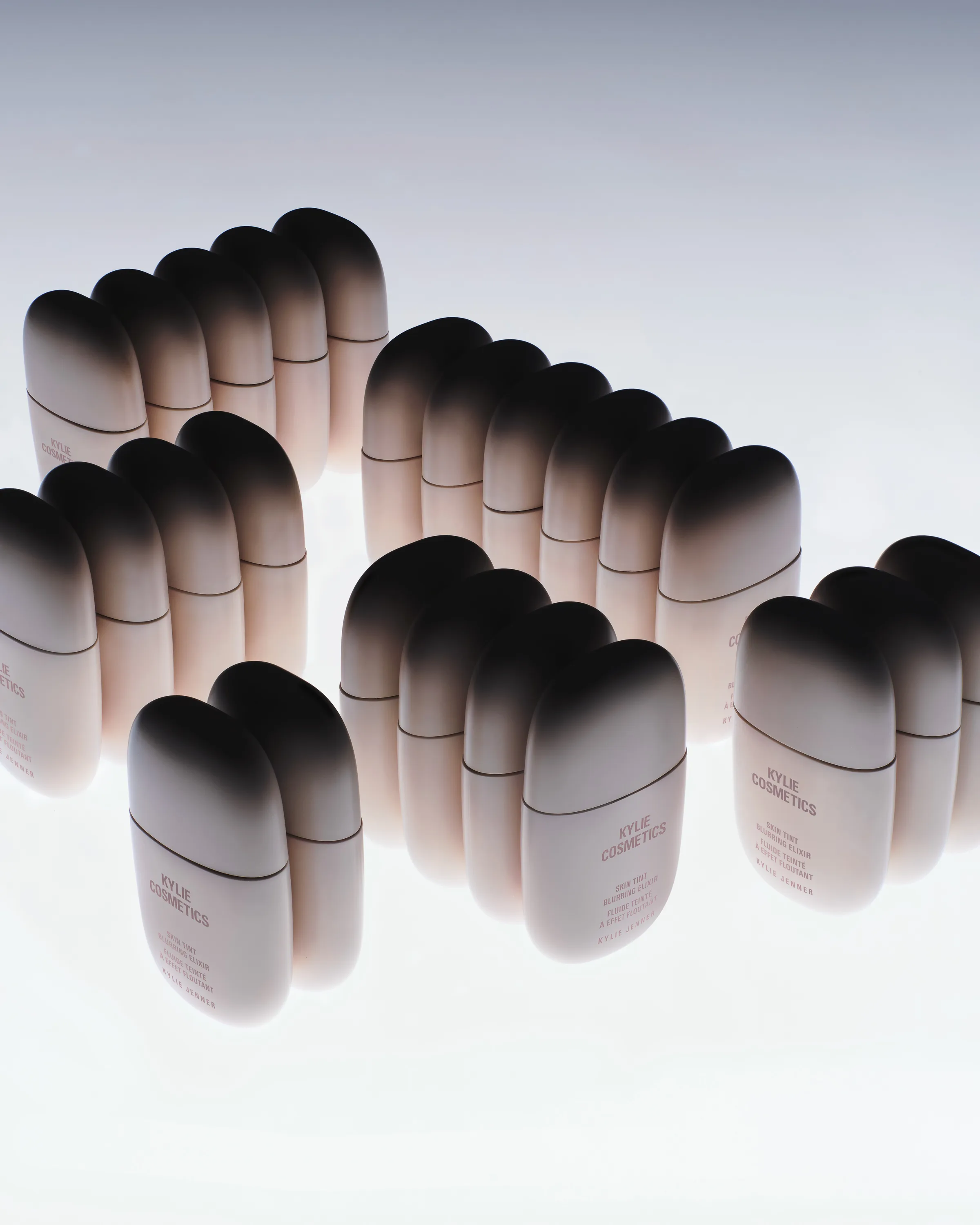 Rows of pale pink skin tint bottles with gradient dark caps, grouped on a glowing white surface, for Kylie Cosmetics.