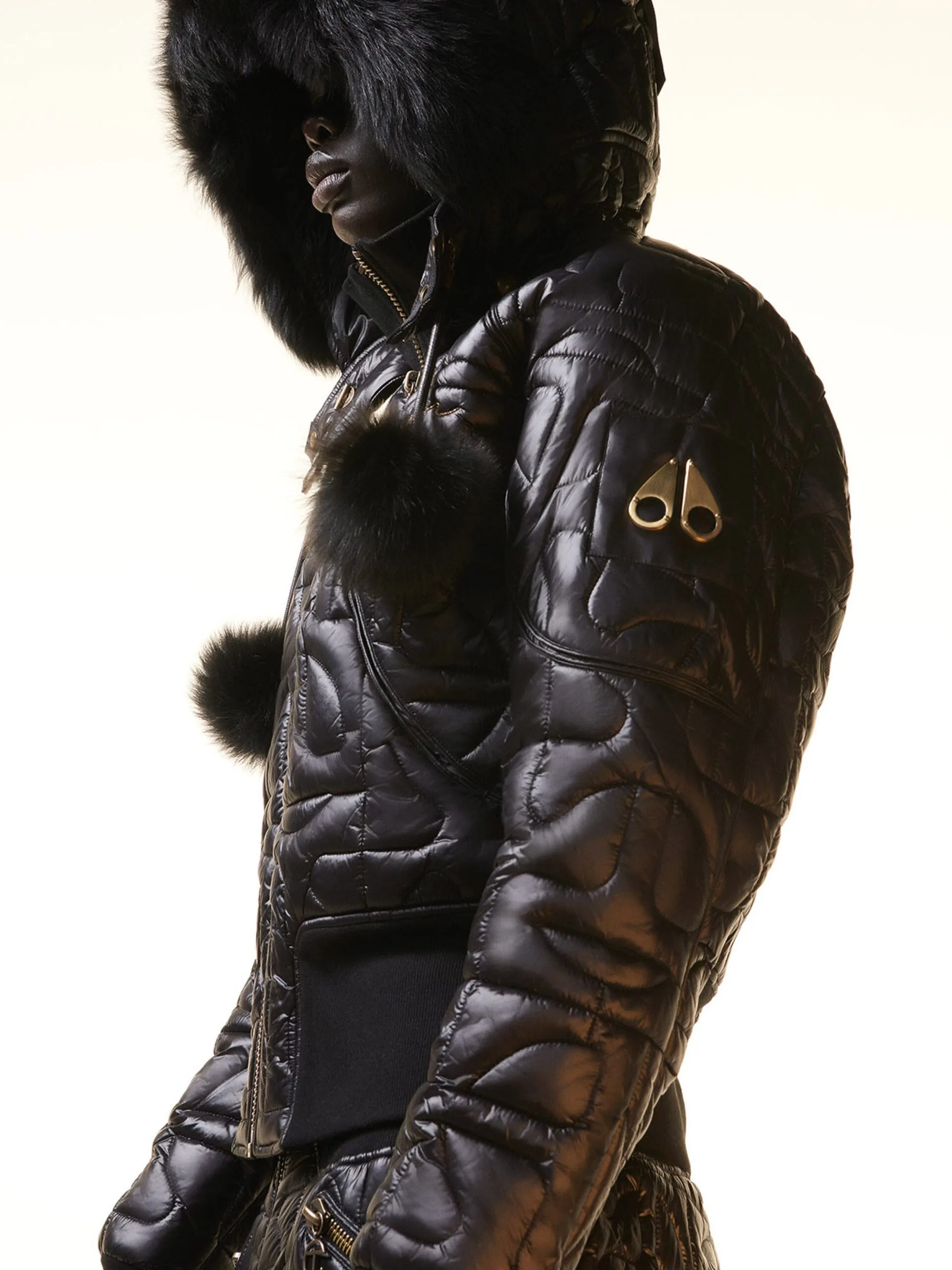 Model in profile wearing a glossy black quilted bomber jacket with fur hood and pom-poms, for Moose Knuckles.