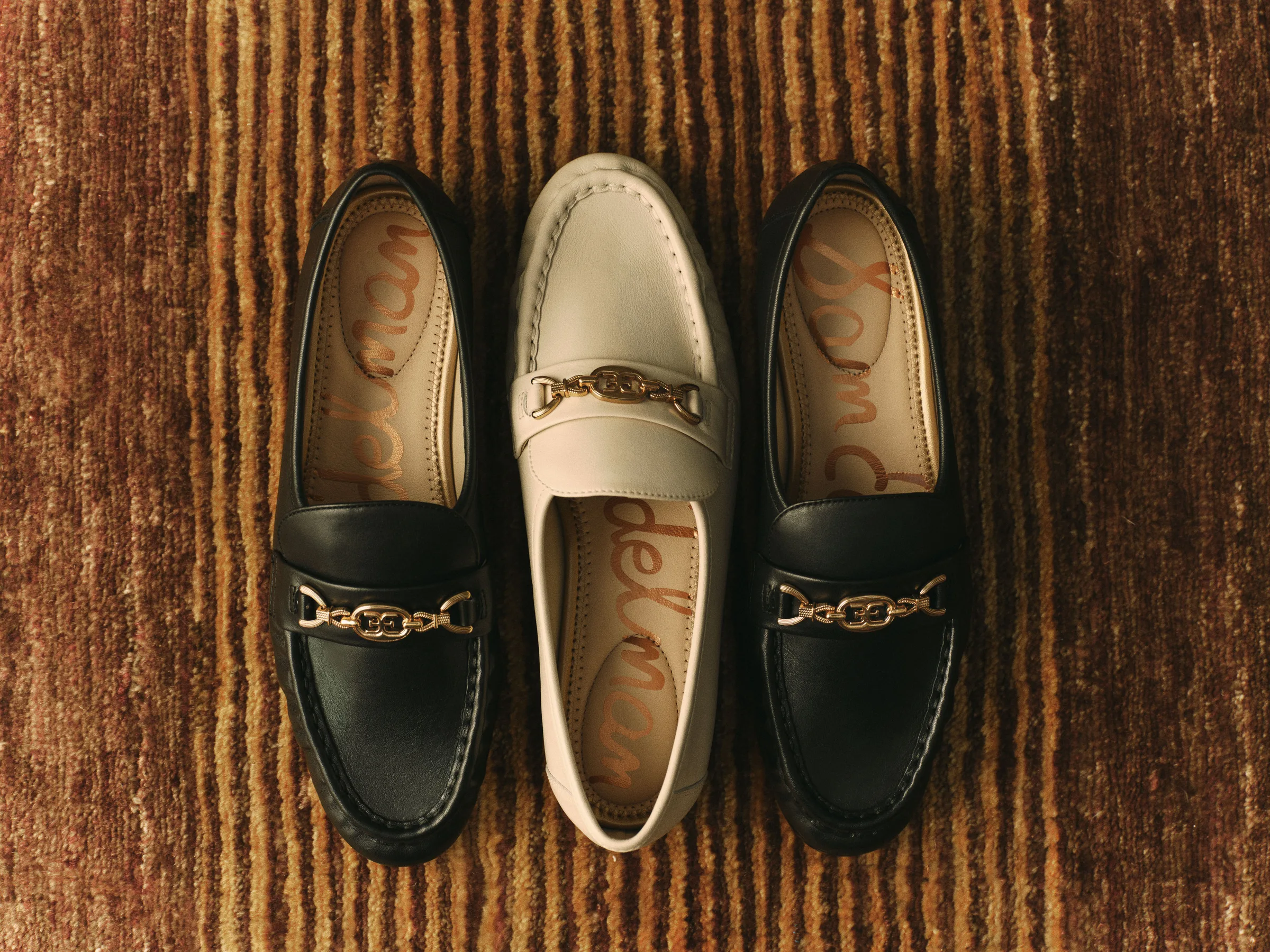 Black and ivory leather loafers with gold chain hardware laid on a striped brown rug, for Sam Edelman.