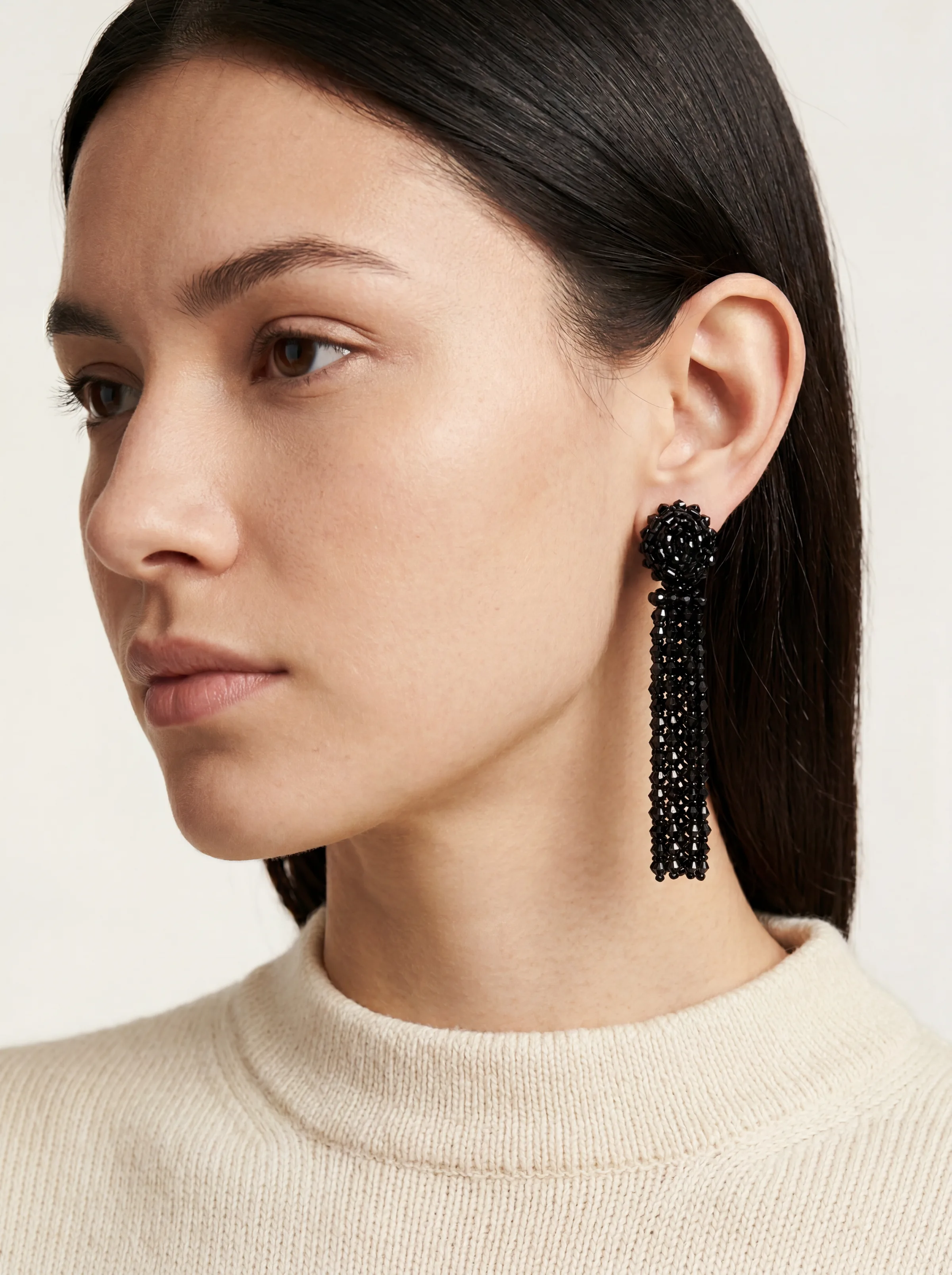 On-model close-up of a black beaded tassel earring worn with a cream knit sweater. Oscar de la Renta AI Concept.