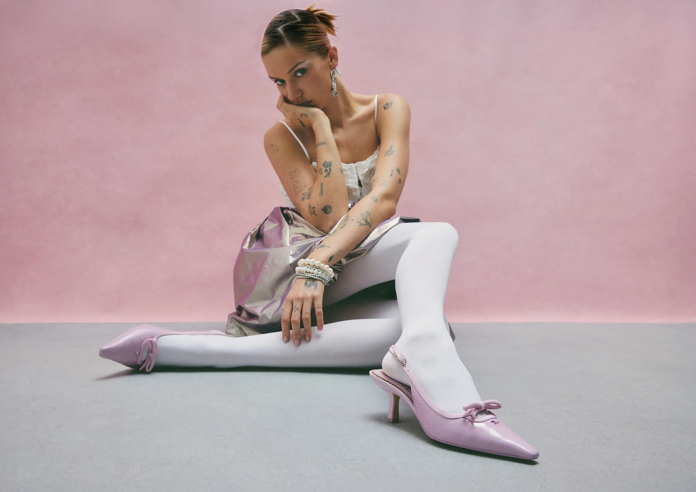 Tattooed model seated on grey floor in white tights and pale orchid bow slingback heels against a pink backdrop, for Sam Edelman.
