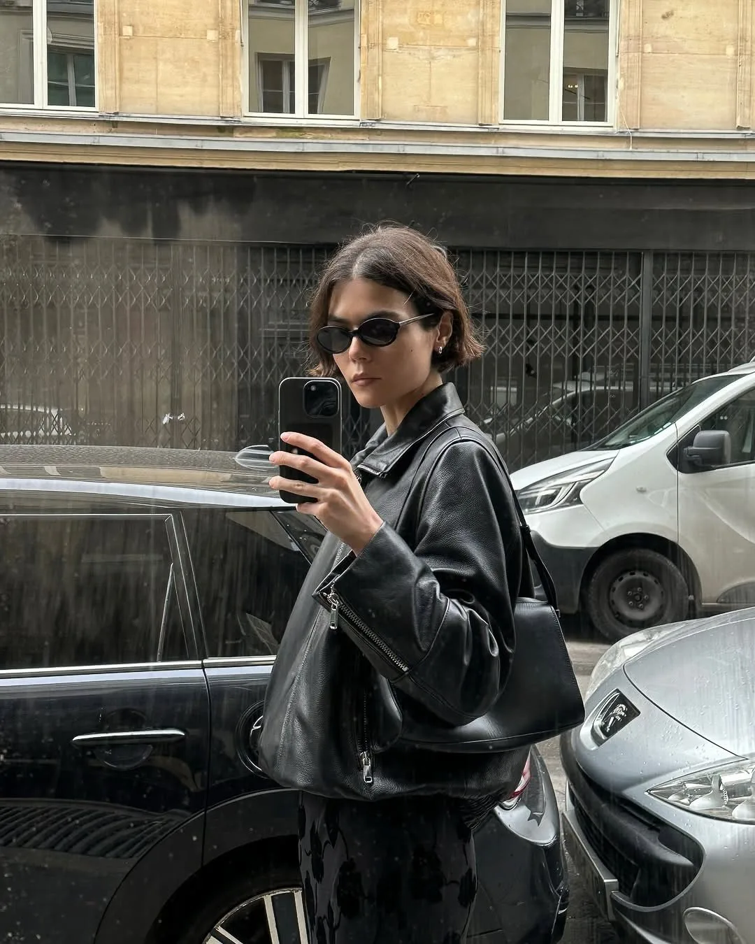 Woman in black leather jacket and sunglasses takes a phone selfie reflected in a parked car on a city street.