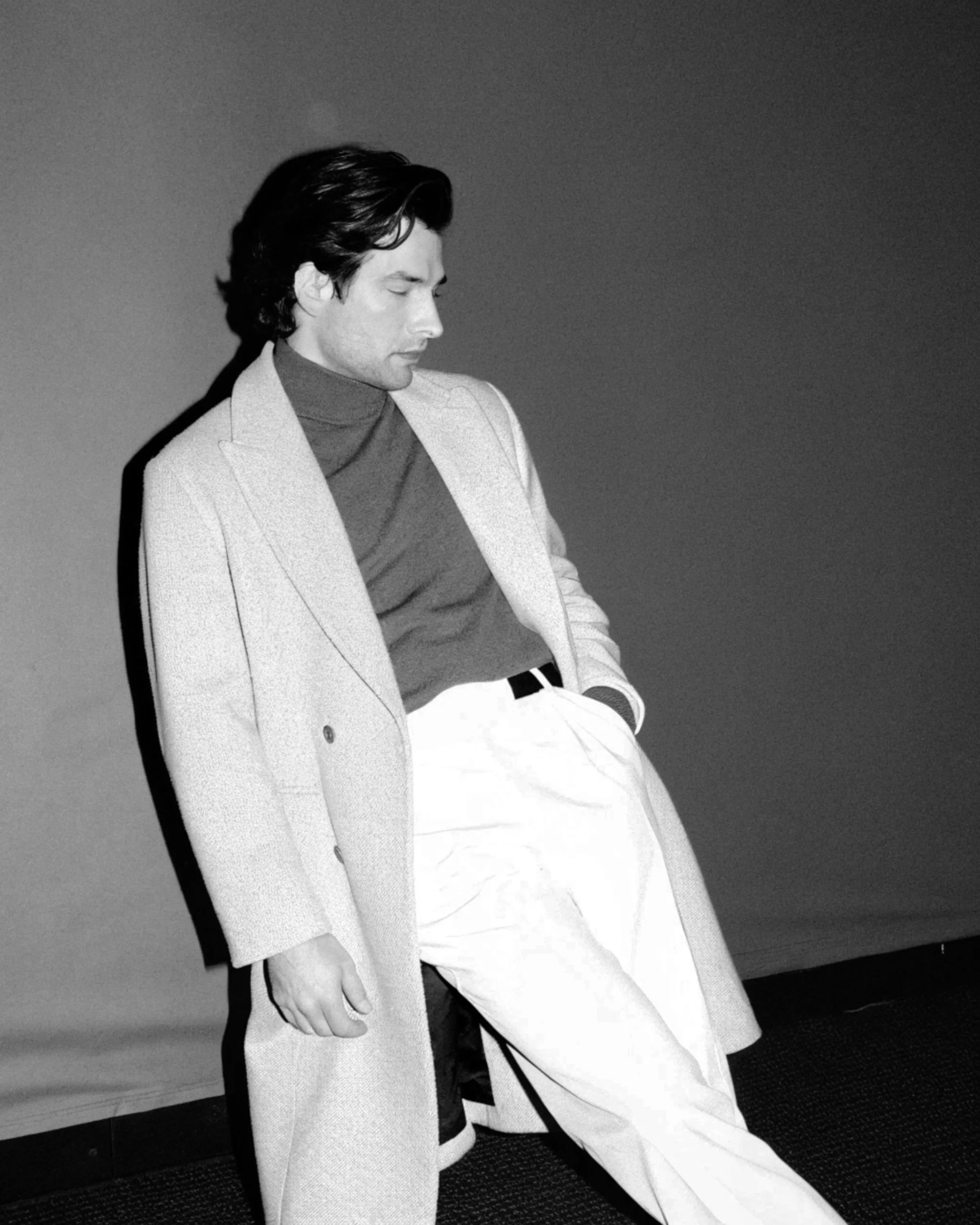 Black-and-white AI image of a man in a long overcoat, turtleneck and wide trousers leaning on a wall. Fursac AI Concept.