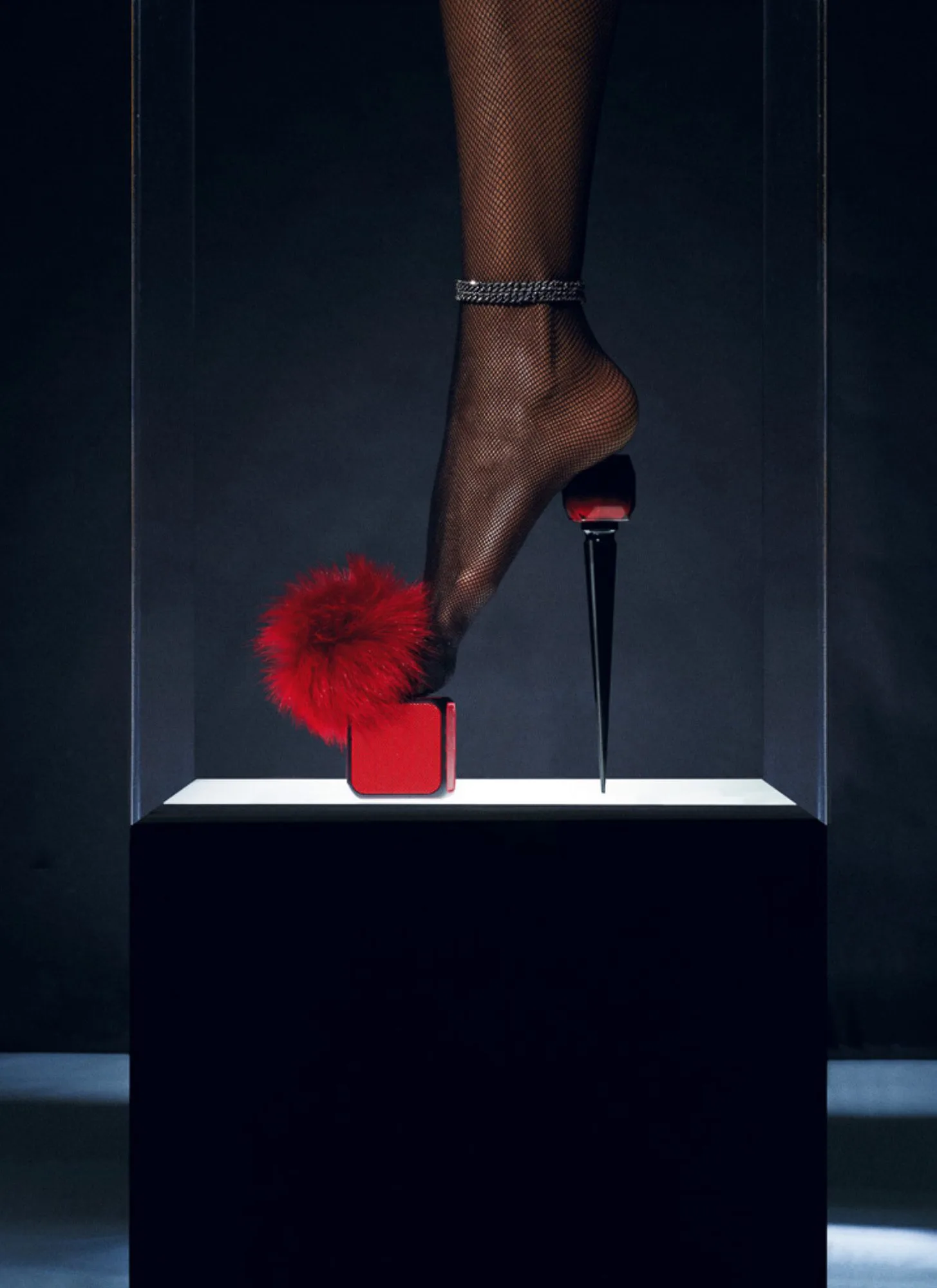 Leg in black fishnet with a jeweled anklet poised on a red cube and black spike heel in a lit glass display case, for Interview Magazine.