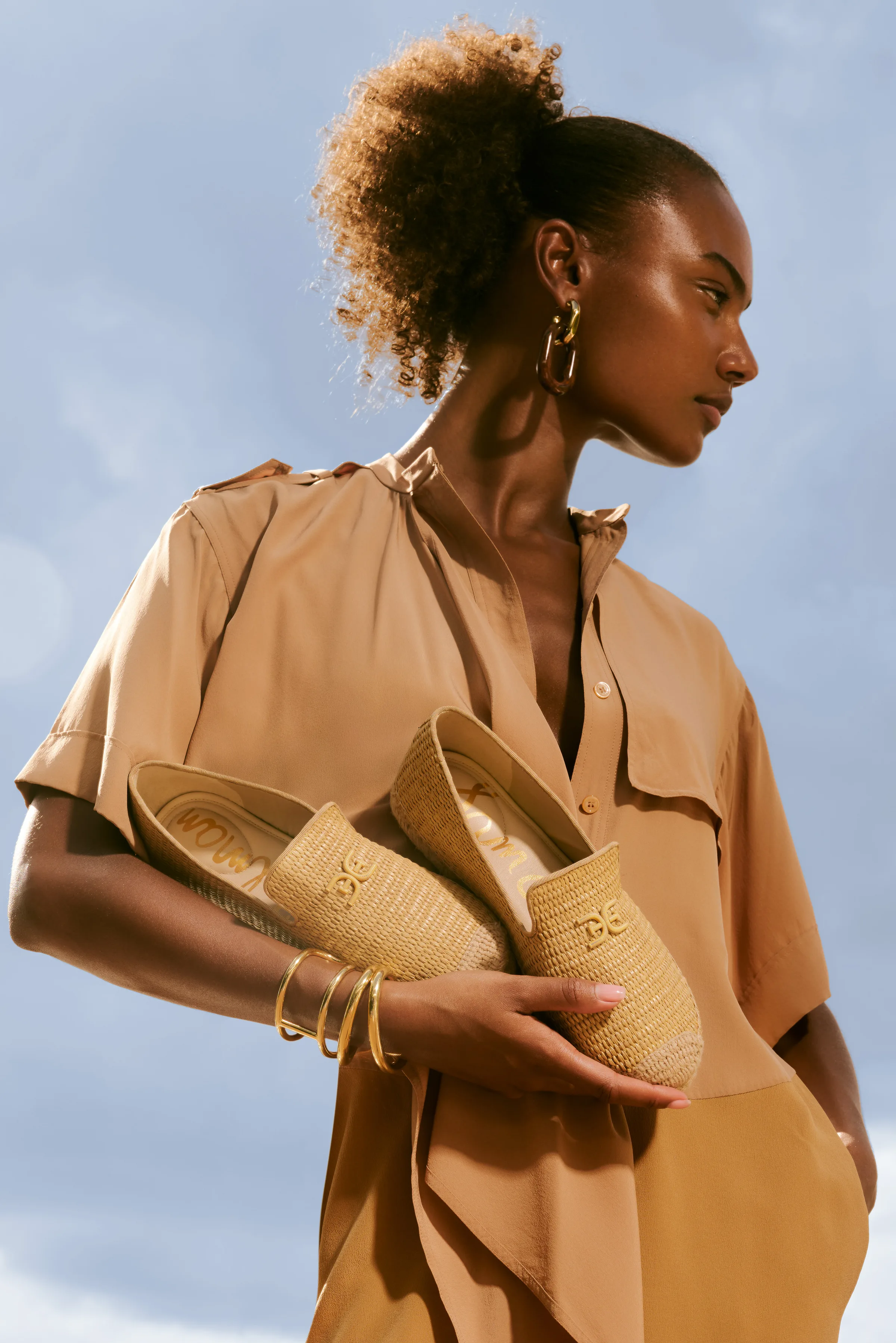Model in a tan shirt dress holding woven raffia espadrille flats with gold logos against a blue sky, for Sam Edelman.
