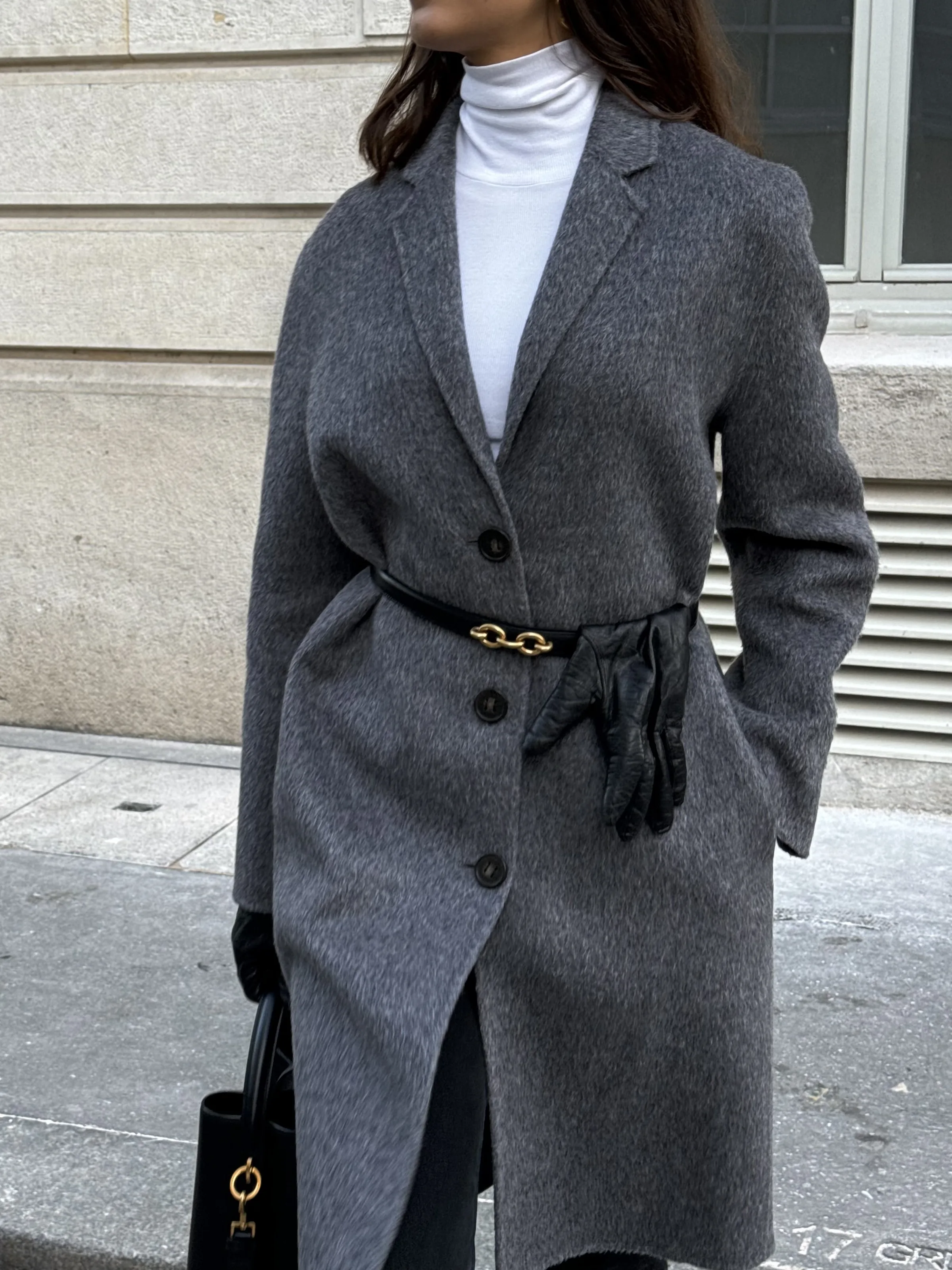 Woman on a city sidewalk in a grey wool coat, white turtleneck, gold-link belt with tucked black gloves and black bag.