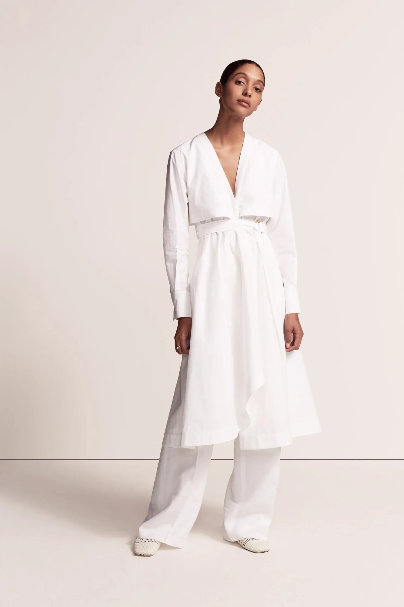 Model in a white belted V-neck shirtdress over wide-leg white trousers against a cream backdrop, for Another Tomorrow.